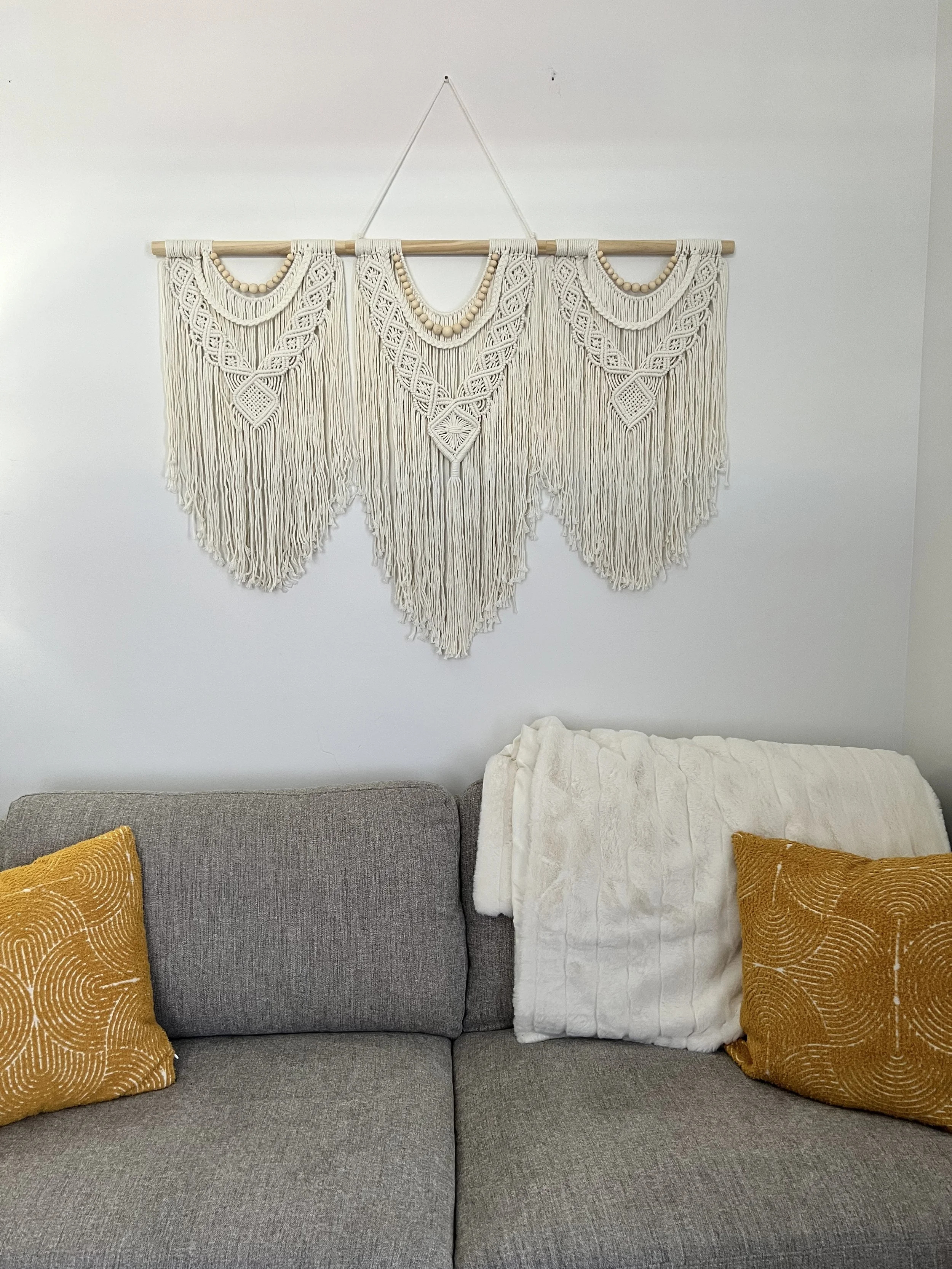 A living room with a gray sofa decorated with two mustard-colored pillows, a white blanket, and a wall hanging made of macramé with intricate patterns and fringe details on a white wall.
