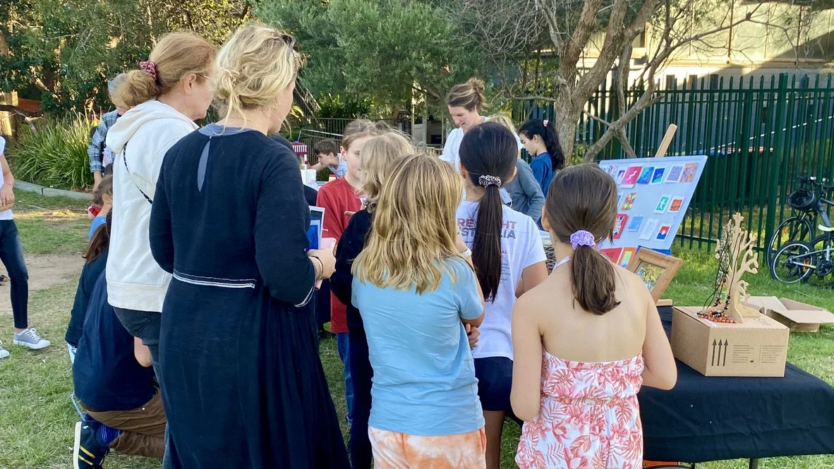 Children and adults gathered around outdoor display boards and tables at a park event.