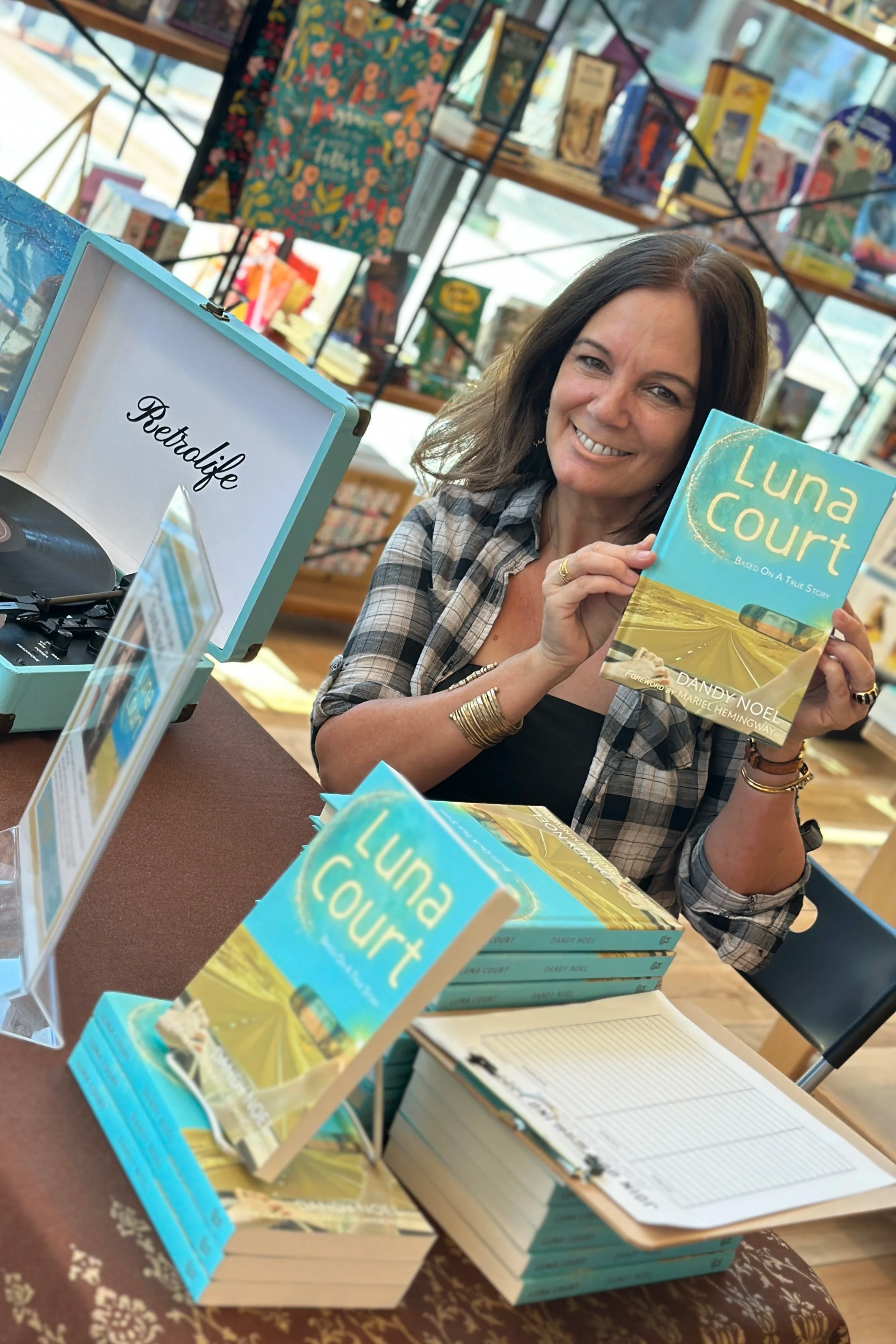 A woman smiling and holding up a book titled "Luna Court" at a book signing event, with more copies of the book and a sign that says "RetraLfe" on the table, inside a bookstore.