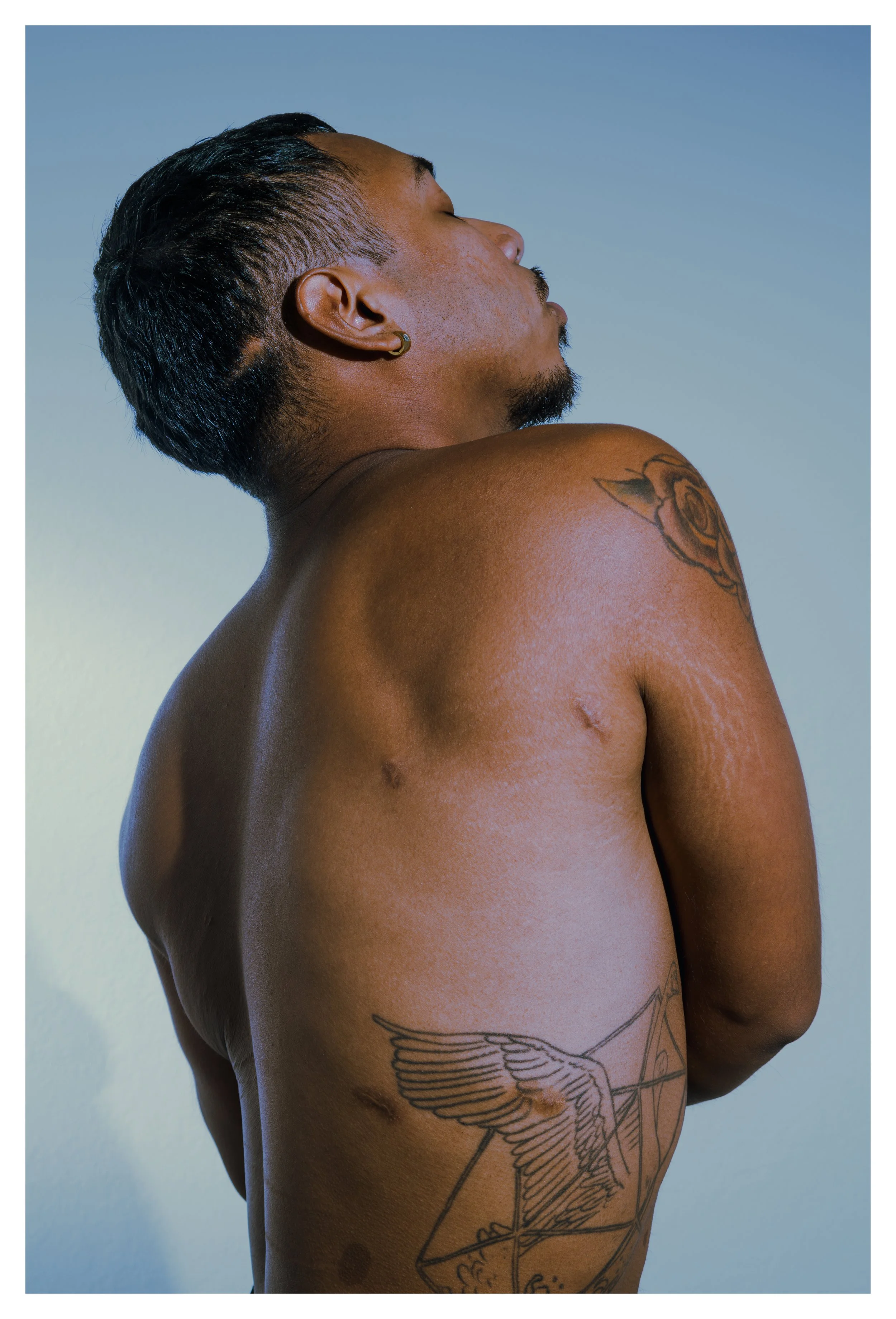 Side profile of a shirtless man with tattoos on his shoulder and side, with short black hair, closed eyes, and a relaxed expression, against a plain background.
