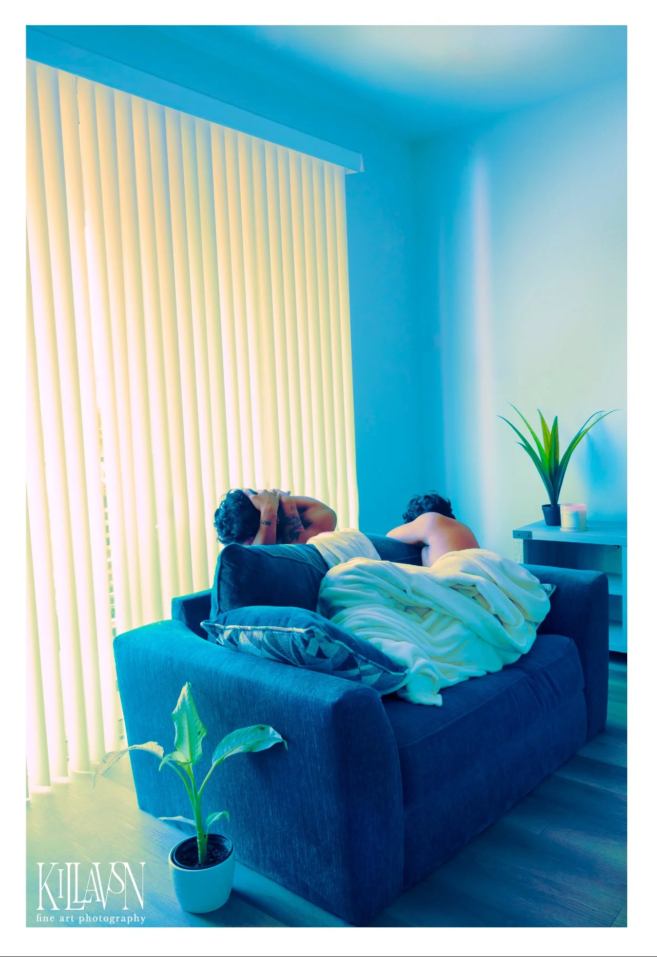 Two people are sleeping on a blue sofa in a room with vertical blinds and a plant on a side table.