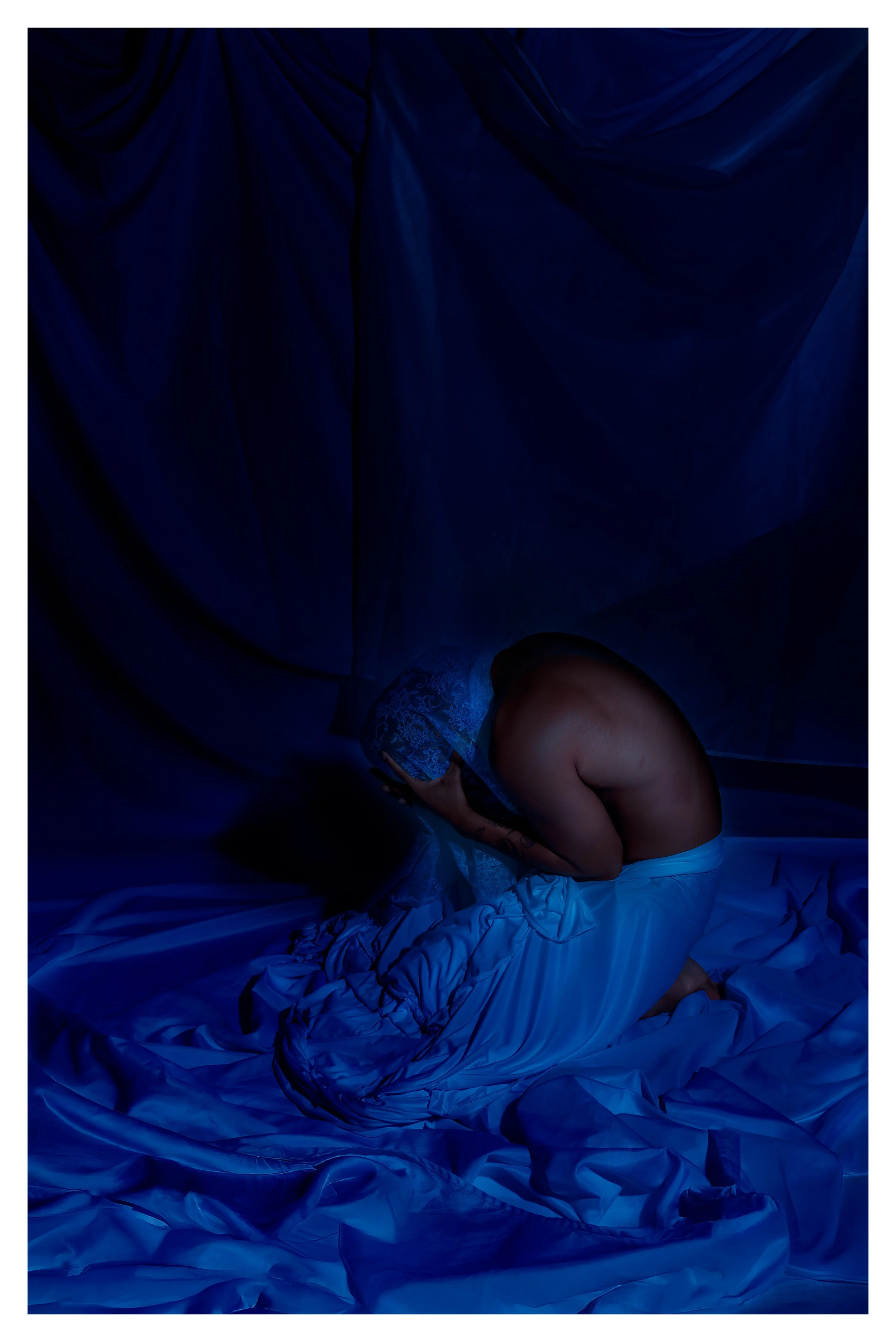 A person kneeling on a bed, covering their face with a bowl, surrounded by blue satin sheets and curtains, illuminated by dim blue lighting.