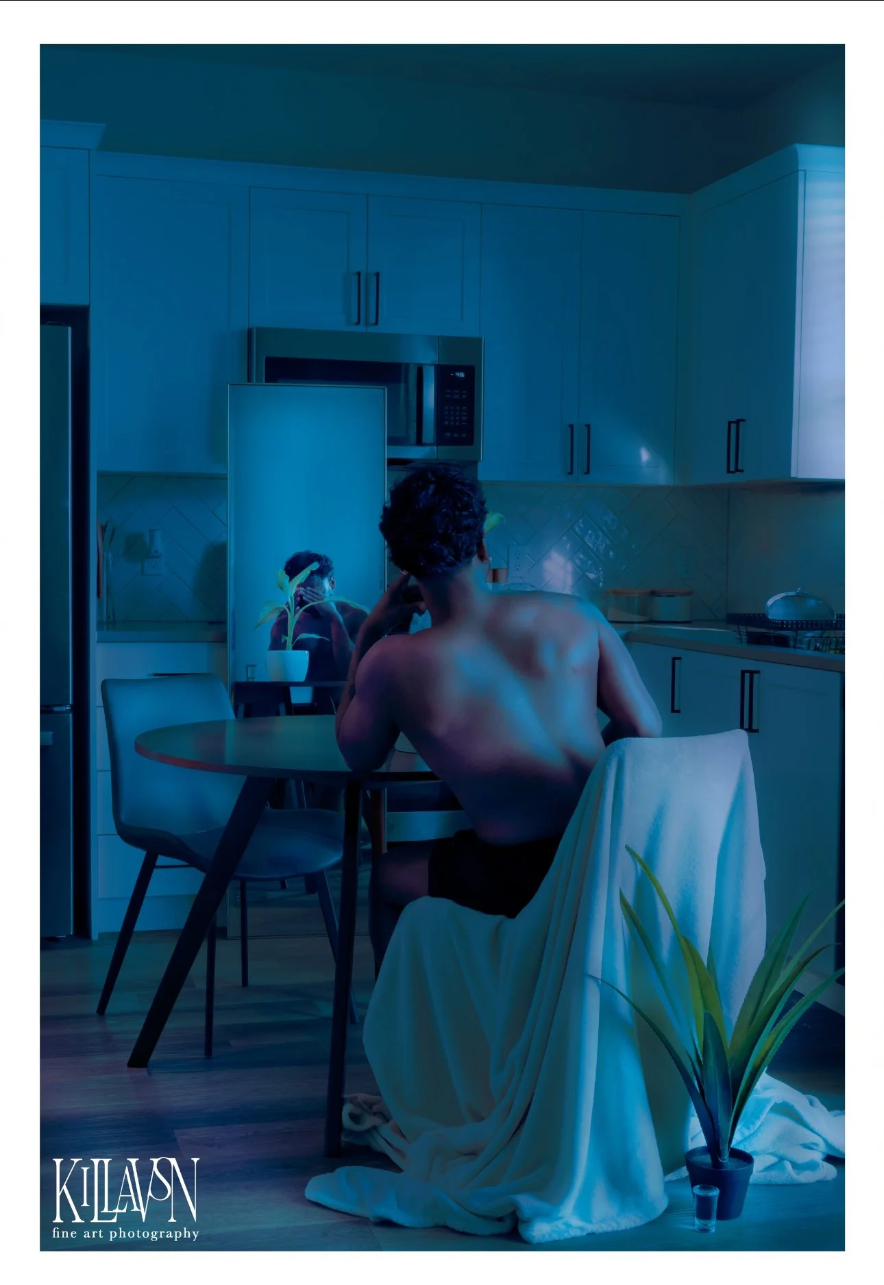 A shirtless person with curly hair sitting at a round dining table in a modern kitchen, with a potted plant in the foreground, illuminated by blue lighting.