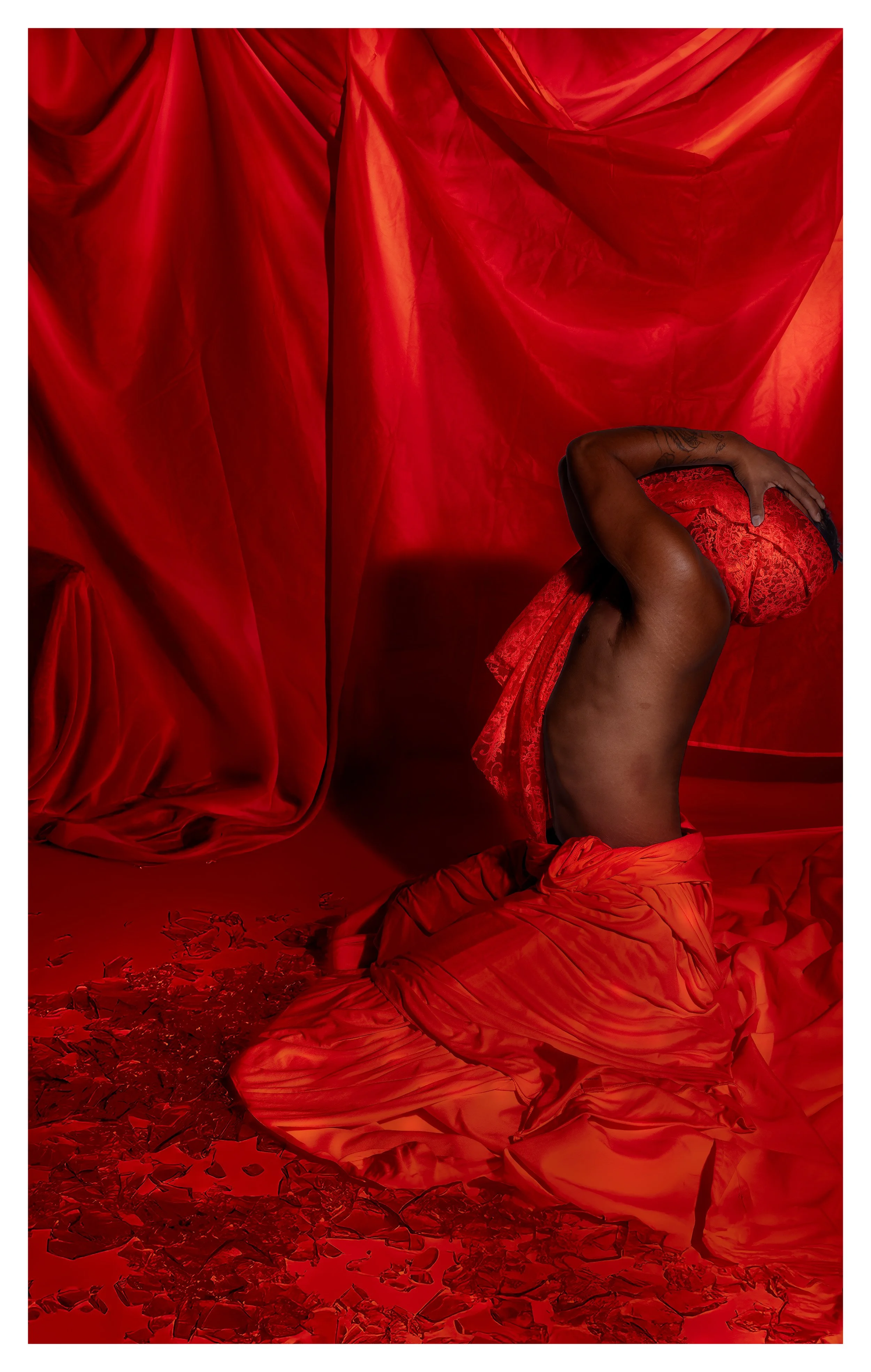 A person wearing a red lace garment covering their head, nude from the waist up, seated on a red fabric surface surrounded by broken glass, with a red draped fabric backdrop.