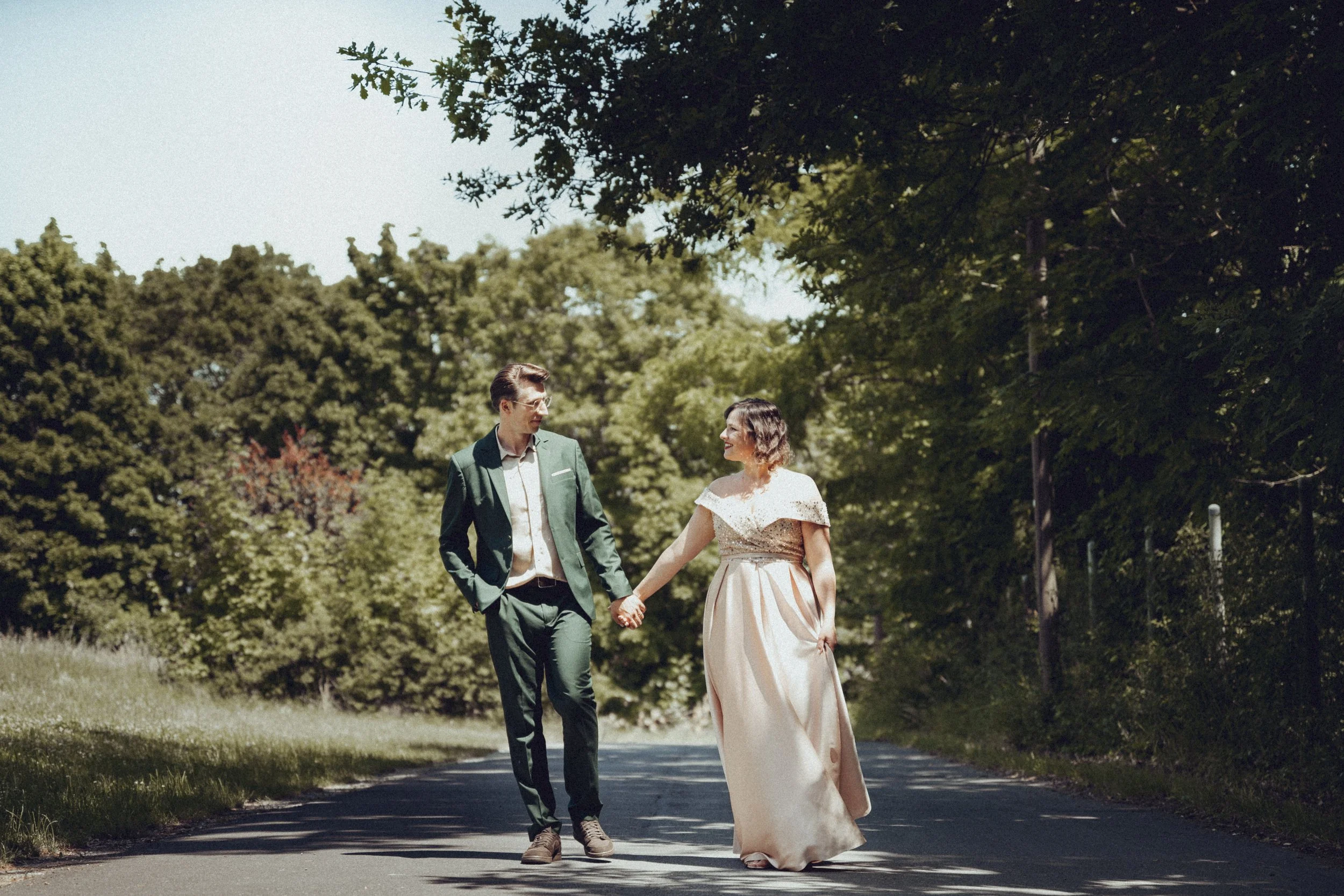Ein Paar in Hochzeitskleidung läuft Hand in Hand auf einer ruhigen Straße umgeben von Bäumen. Er trägt einen dunkelgrünen Anzug, ihr Kleid ist in einem zarten Rosa gehalten. 