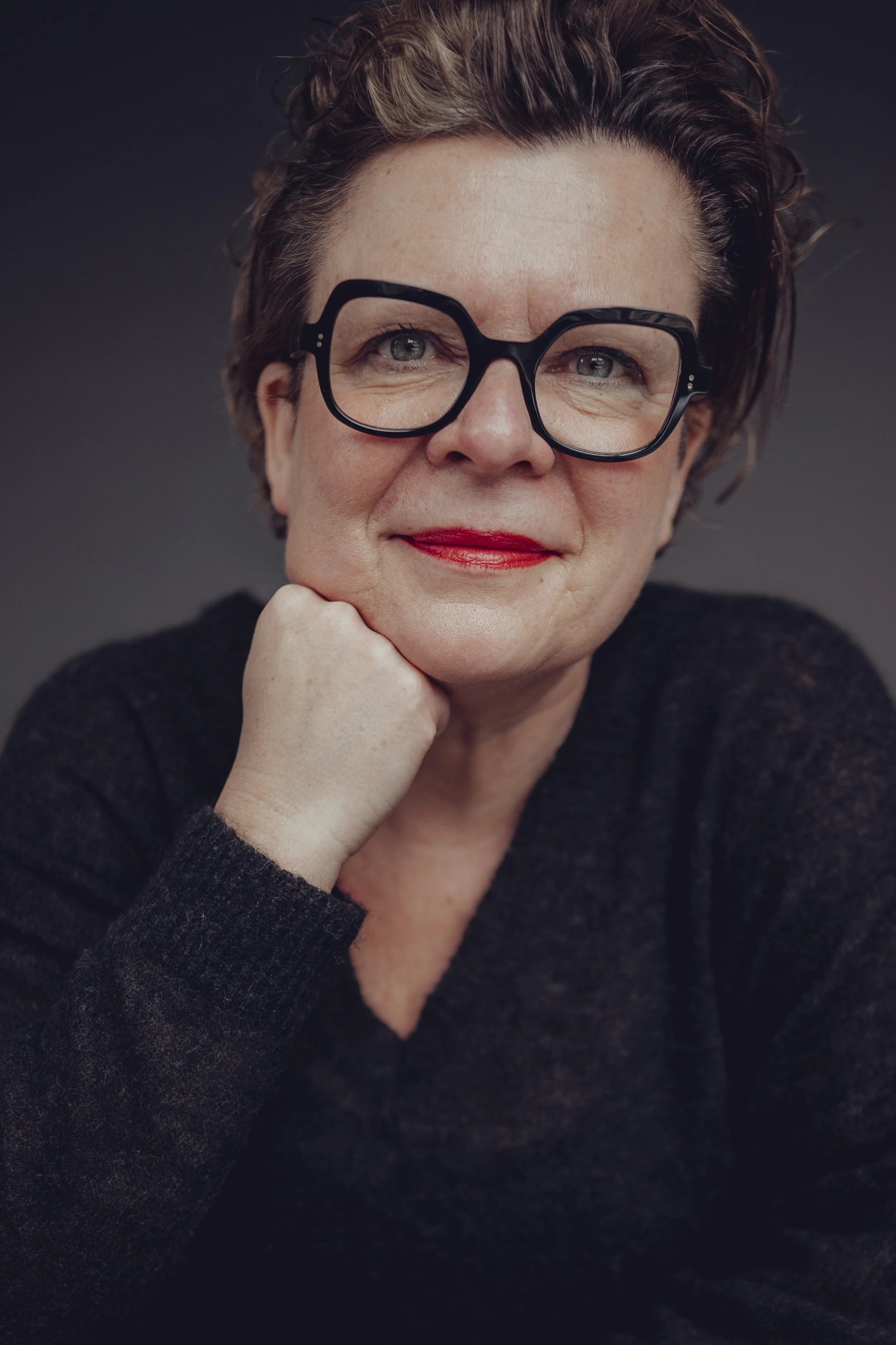 Porträt einer Frau mit Kurzhaarfrisur, Brille und rotem Lippenstift, die lächelnd die Hand an ihr Kinn stützt.