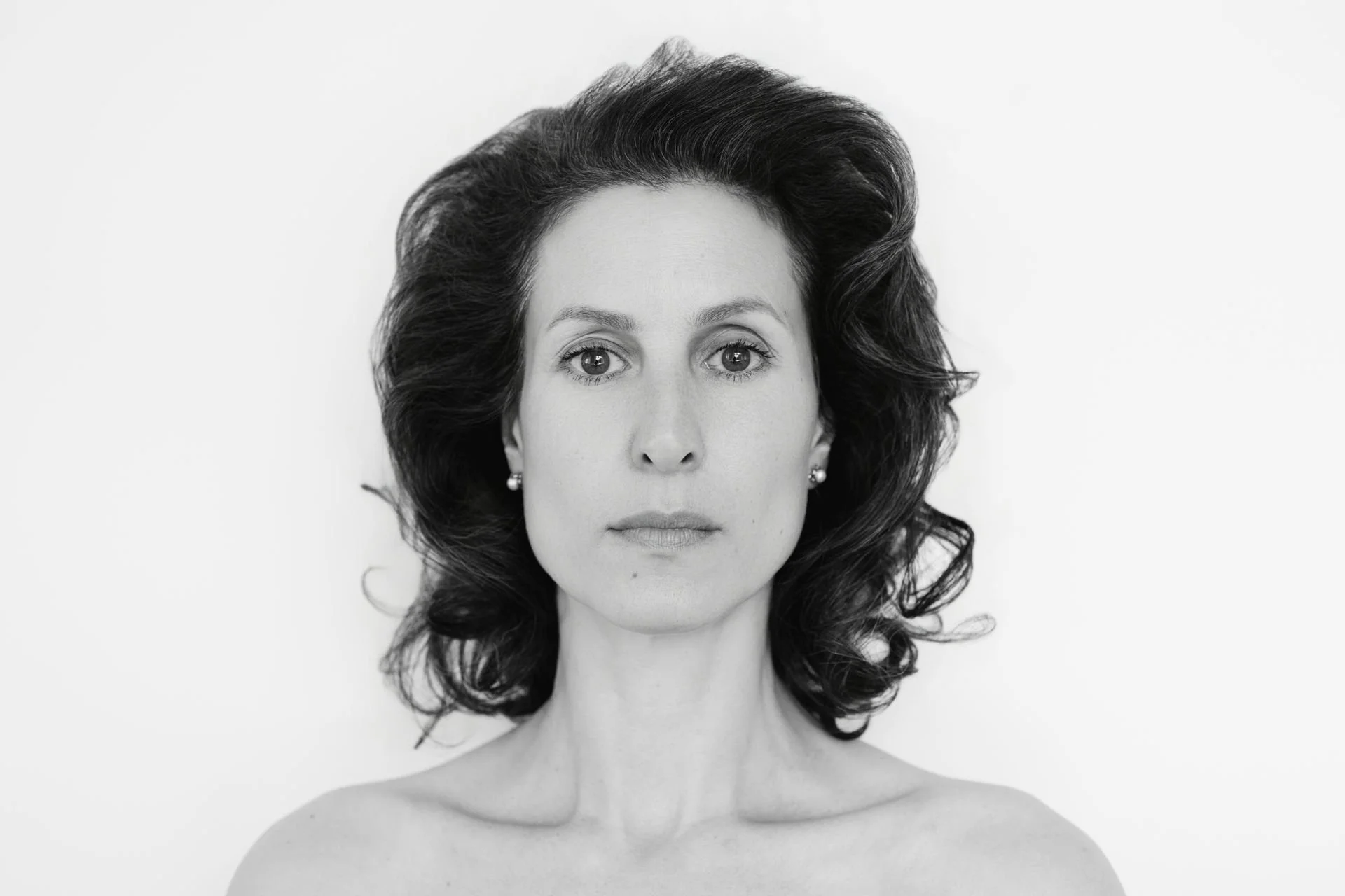 Schwarz-Weiß-Fotografie eines Frauenporträts mit Blick direkt in die Kamera, dunkle, voluminöse Haare, schulterfrei, dezenten Ohrringen, neutraler Gesichtsausdruck, Hintergrund hell und unscharf.