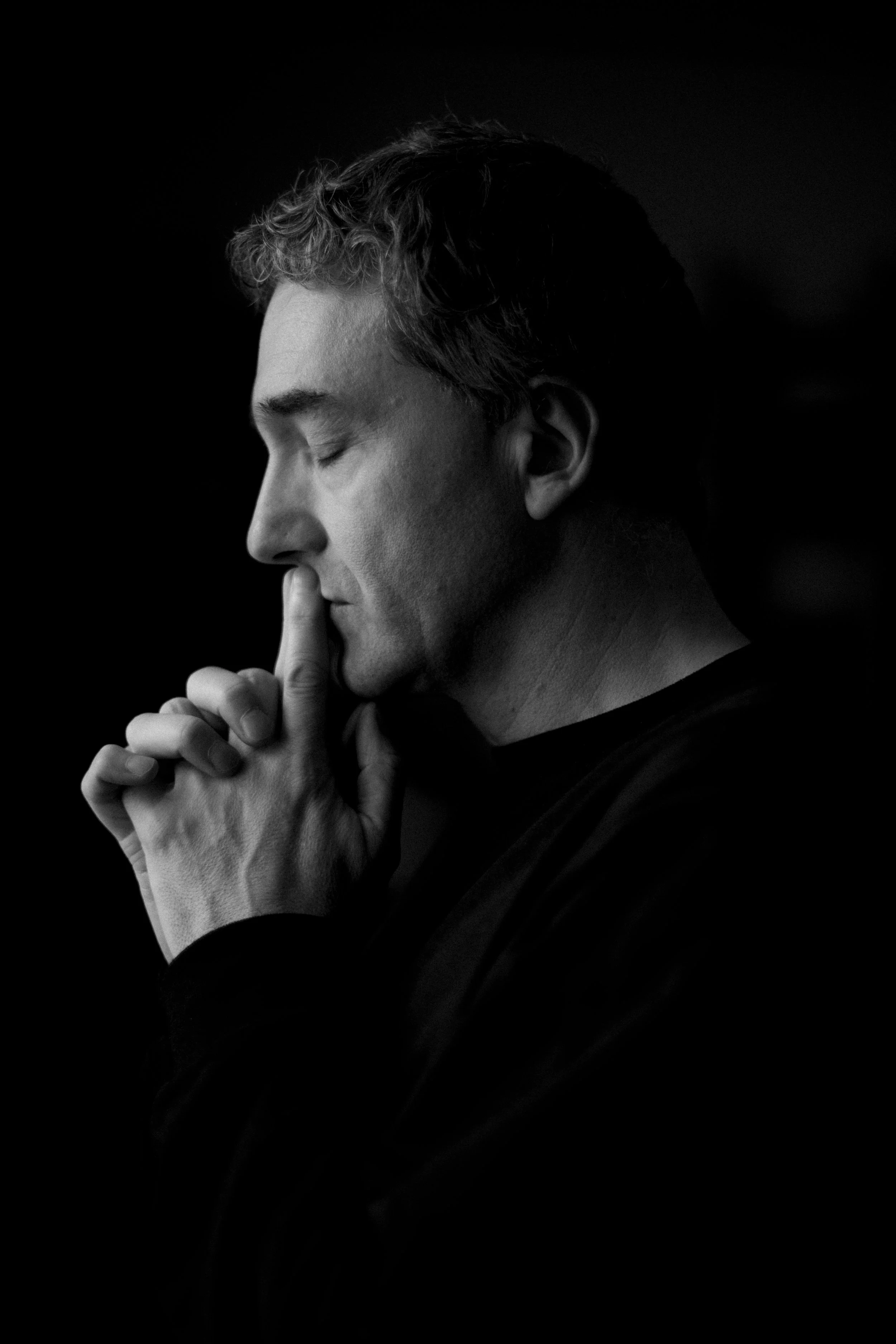 Schwarz-weiß-Foto eines Mannes mit geschlossenen Augen, der die Hände vor den Lippen hält und den Finger auf die Lippen legt, in einer nachdenklichen Pose.