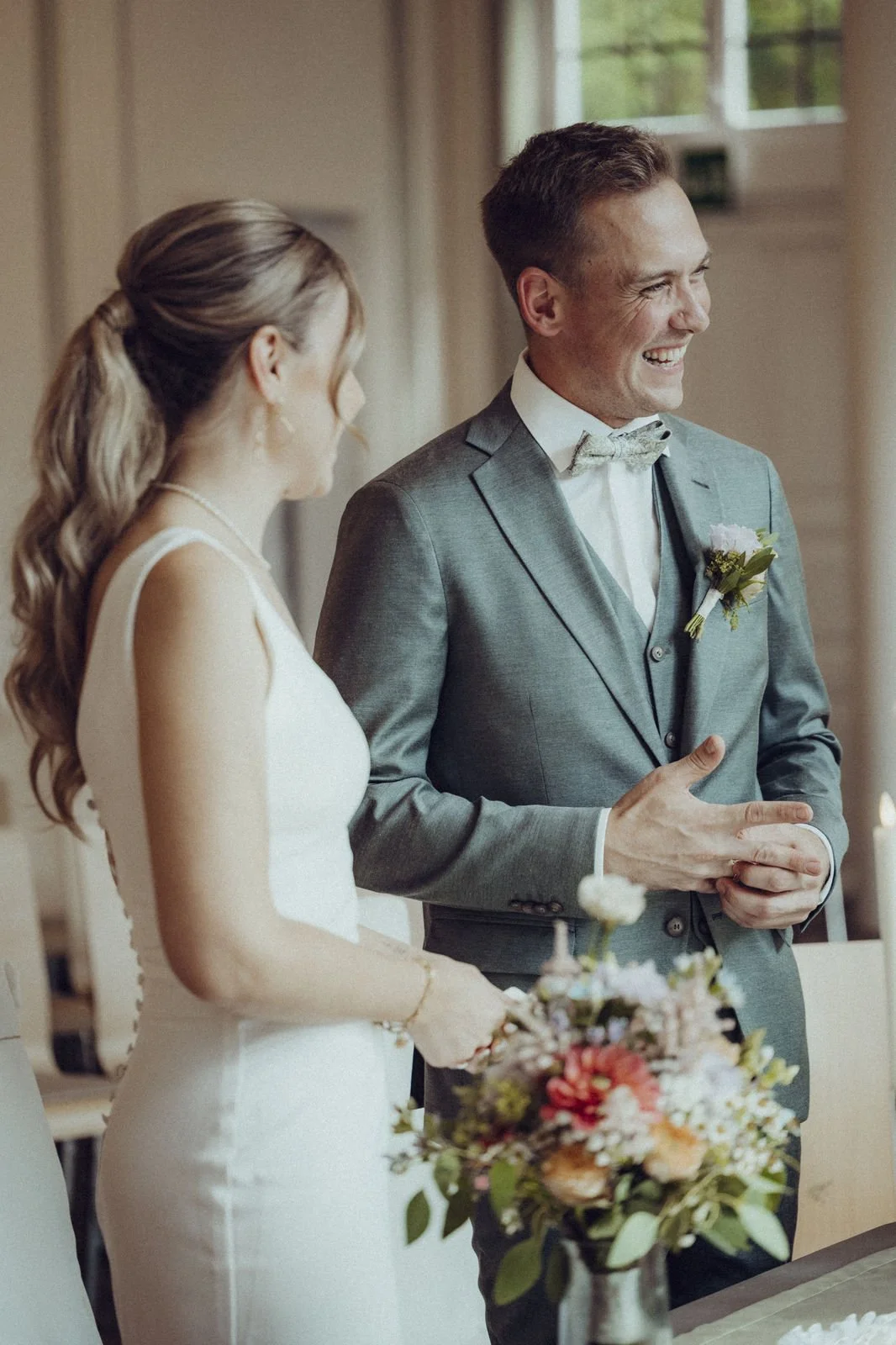 Ein glückliches Brautpaar bei ihrer Hochzeit, der Bräutigam trägt einen grauen Anzug mit Fliege, die Braut hält einen Blumenstrauß.