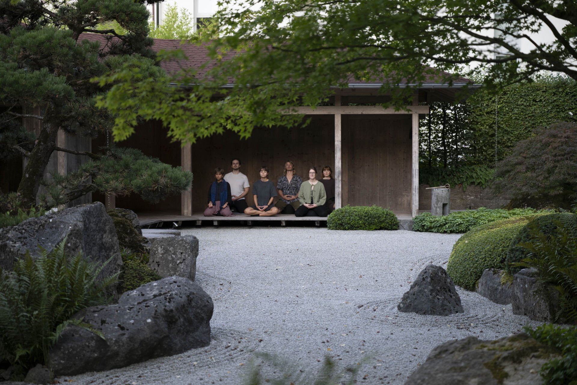 Menschen sitzen im Schneidersitz in einem Holzzelt in einem japanischen Garten, umgeben von Steinen, Büschen und Bäumen.
