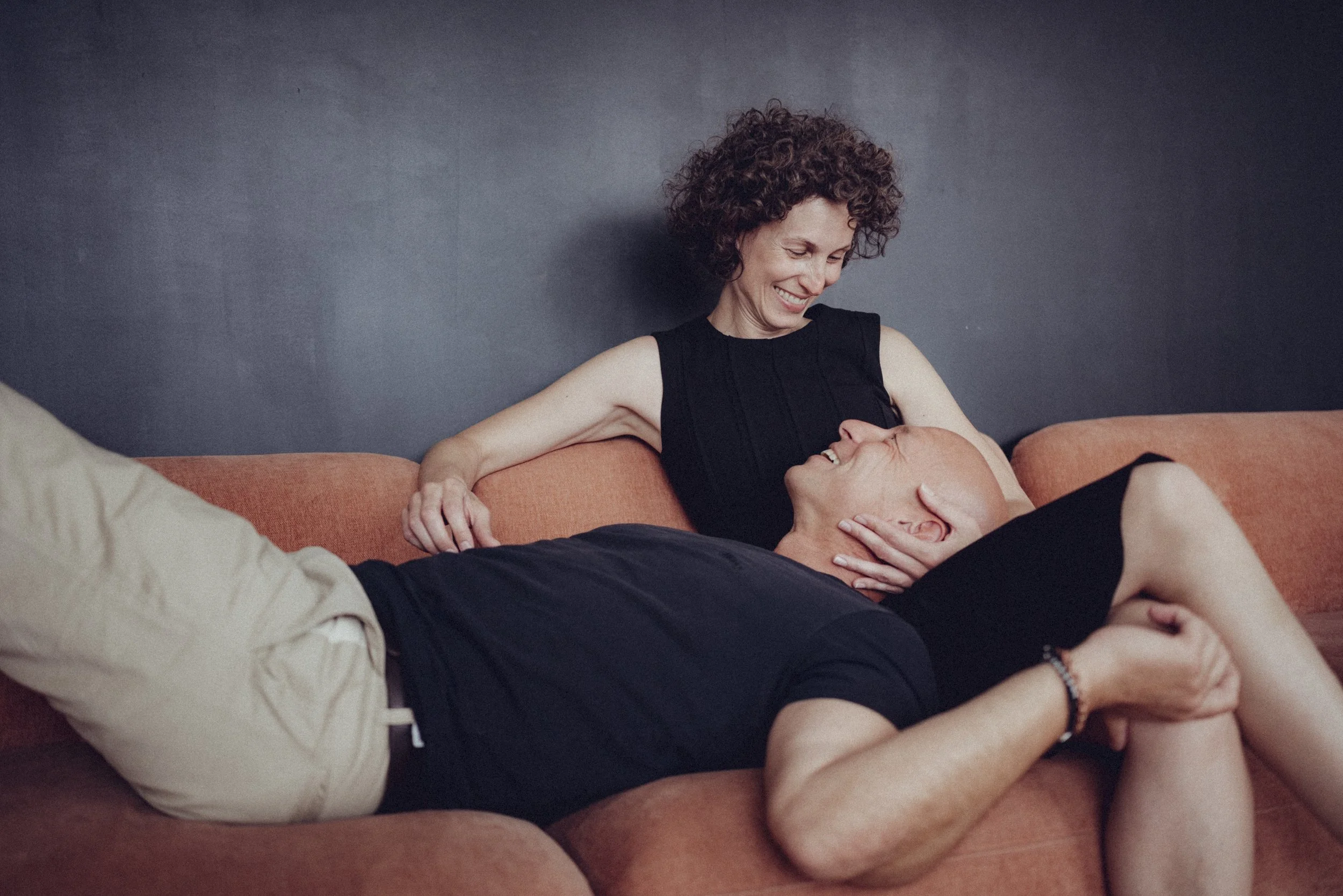 Ein lachendes Paar, das auf einem Sofa sitzt, die Frau hält den Kopf des Mannes, während sie miteinander lachen.