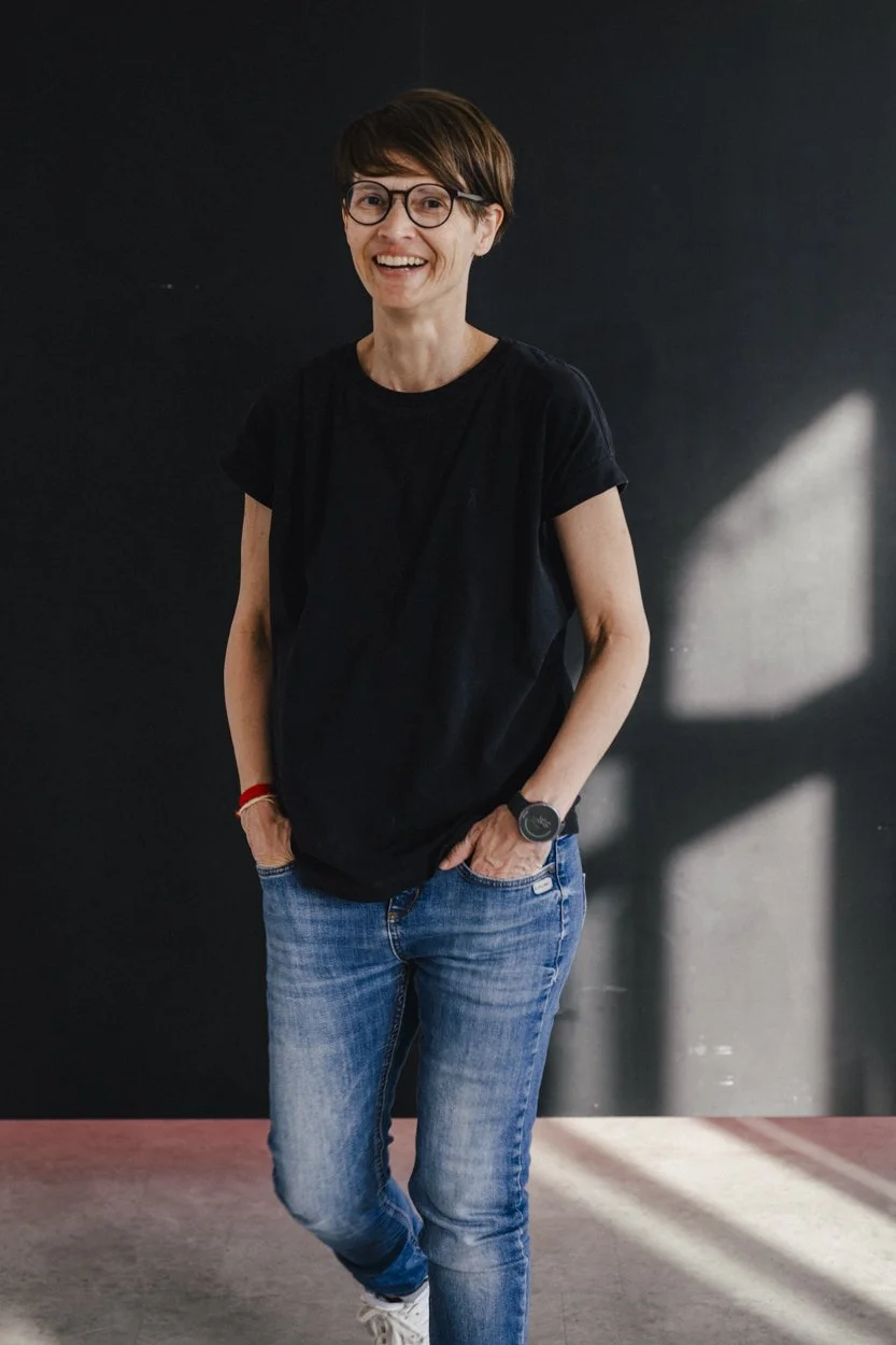 Eine lächelnde Frau mit Kurzhaar, Brille und schwarzem T-Shirt, die auf einer Fläche mit einem dunklen Hintergrund steht.