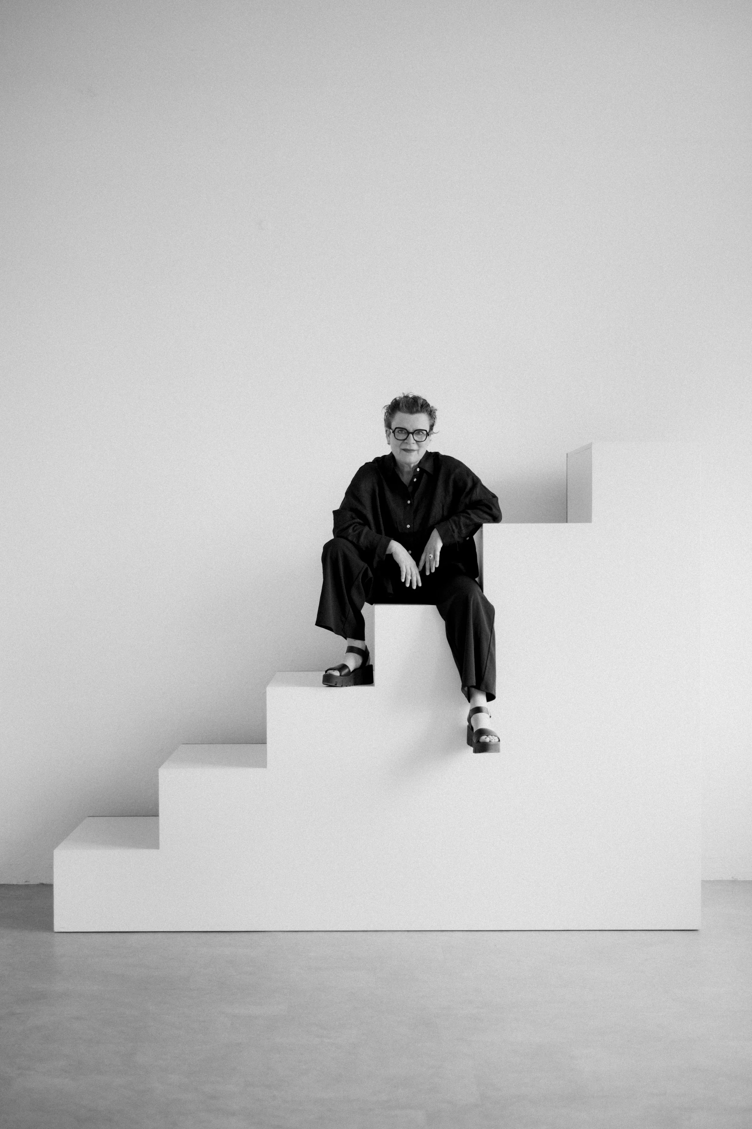 Schwarze und weiße Fotografie einer Frau, die auf weißen Treppen sitzt, mit einem minimalistischen Hintergrund.