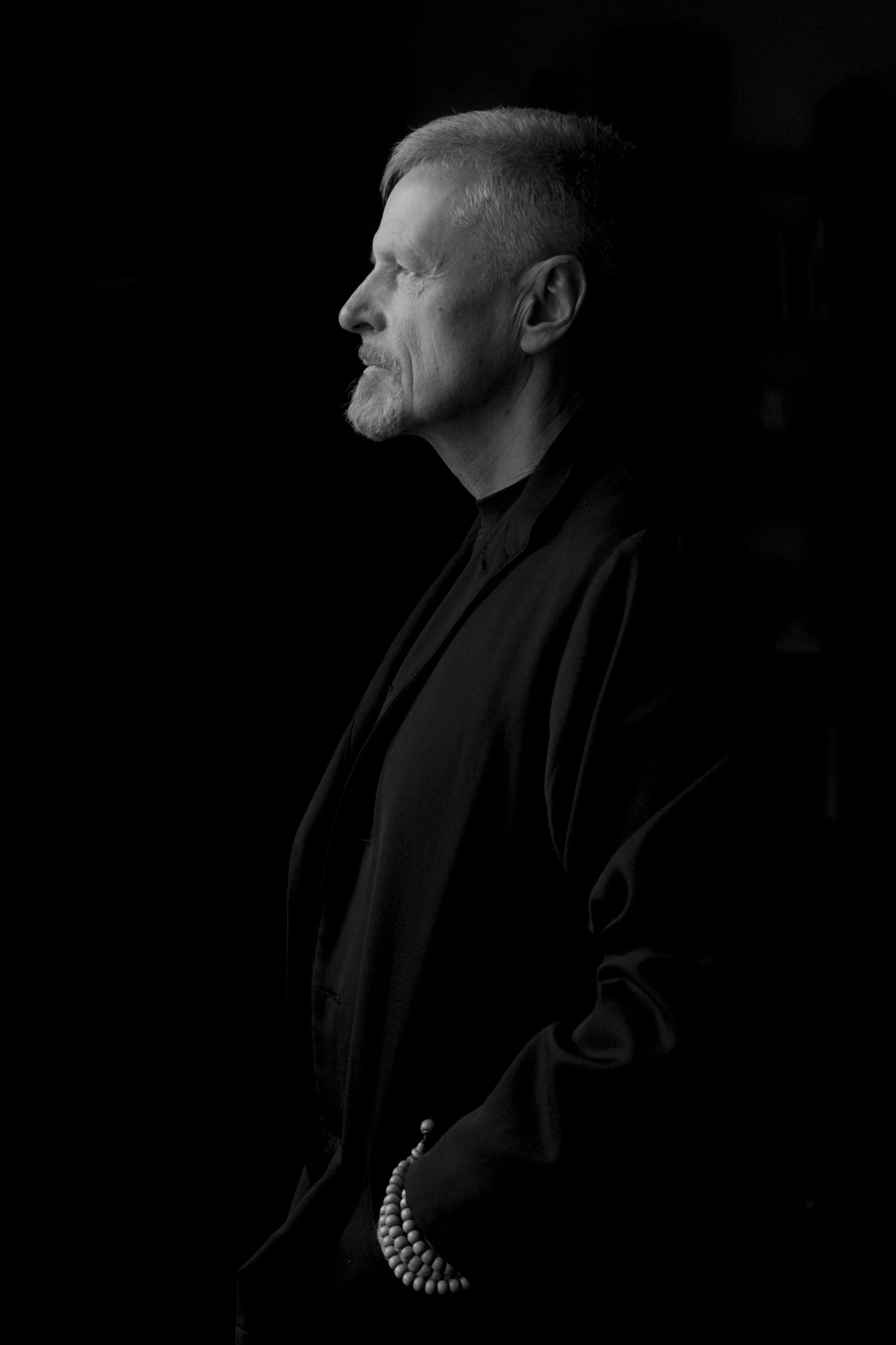 Schwarz-weiß-Fotografie eines älteren Mannes im Profil, mit zerzaustem Haar und einem kurzen Bart, getragen in dunkler Kleidung, mit einer Hand, die eine Perlenkette hält.