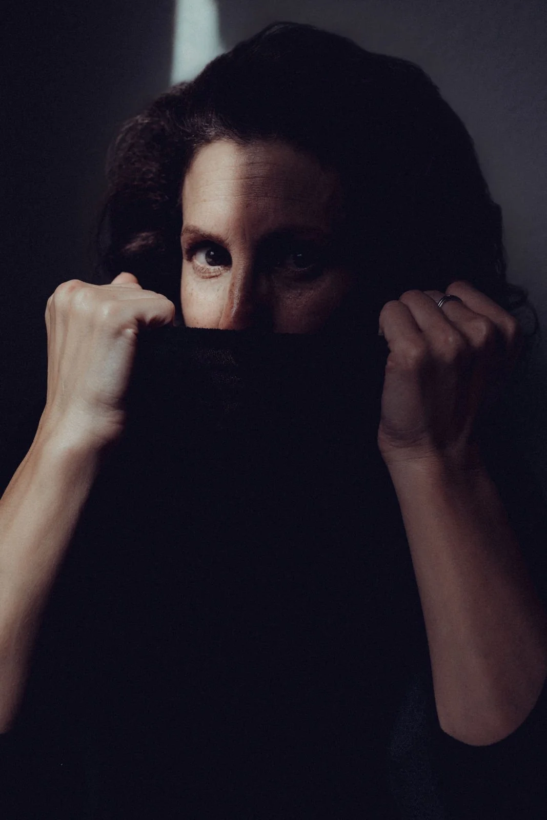 Person, die einen schwarzen Schal oder Pullover trägt, das Gesicht teilweise verdeckt, nur die Augen sichtbar, in einem dunklen Raum.