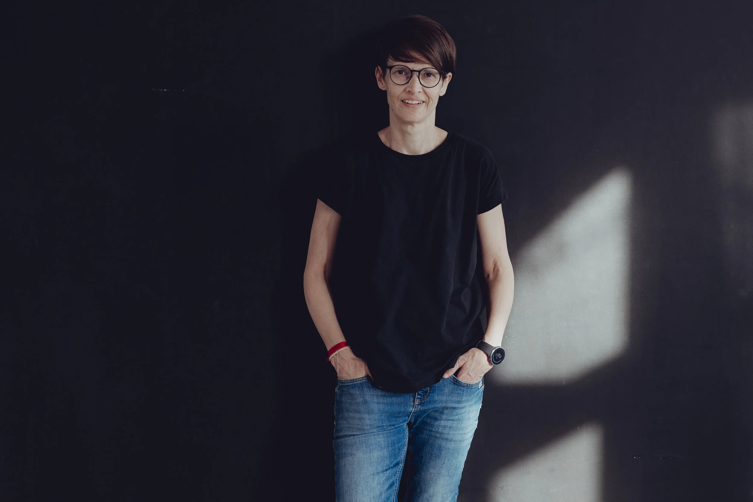 Eine Frau steht angelehnt an eine dunkle Wand. Sie trägt kurze Haare und eine Brille. Die Hände hat sie in den Hosentaschen und trägt ein schwarzes T-shirt, sowie eine blaue Jeans.