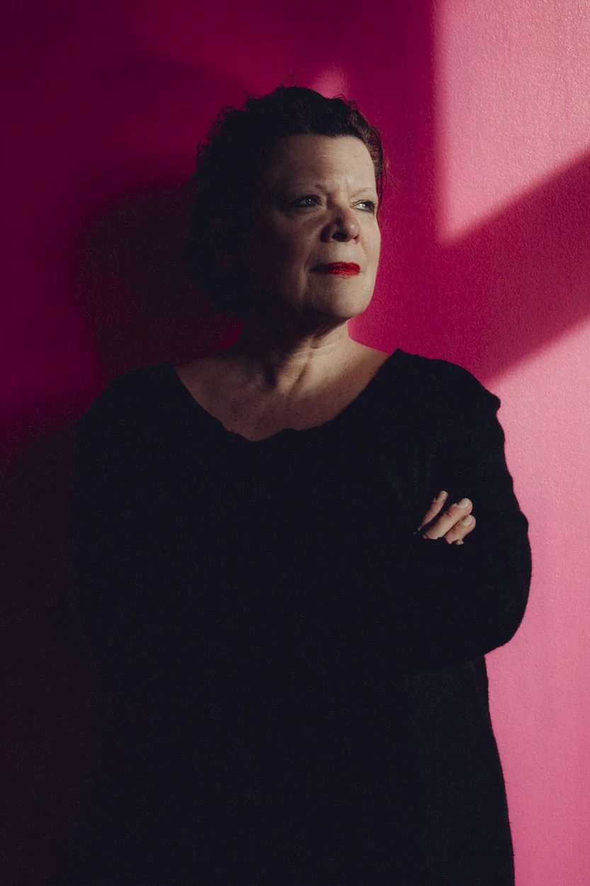 Porträt einer Frau mit kurzen dunklen Haaren vor einer pinkfarbenen Wand, die in den Schatten fällt. Sie trägt ein schwarzes Oberteil, hat rote Lippen und hält die Arme vor der Brust.