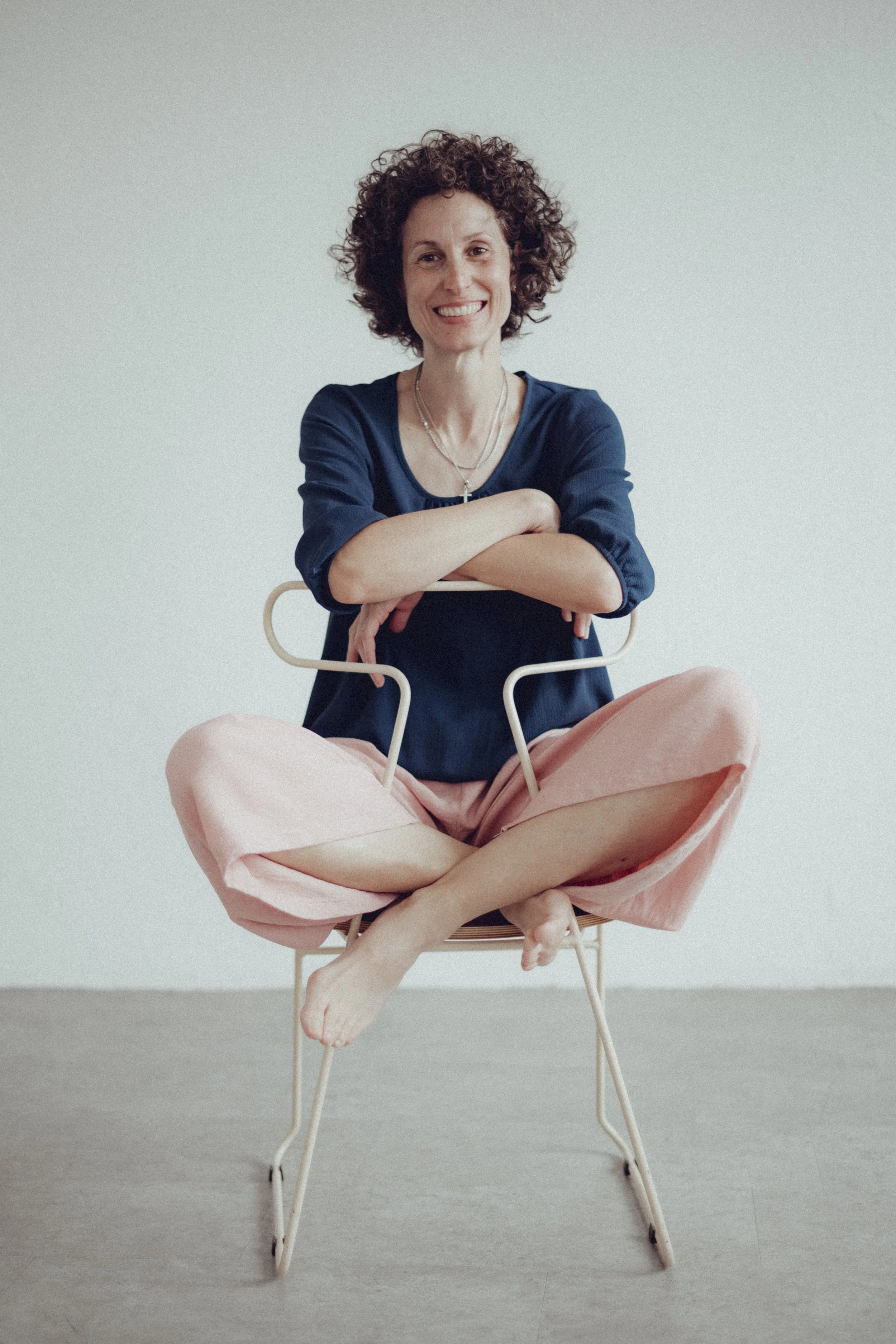 Frau sitzt lachend auf einem Stuhl, die Beine gekreuzt, Hände auf den Armen des Stuhls, trägt eine dunkelblaue Bluse und weiche rosa Hosen, vor einem weißen Hintergrund.
