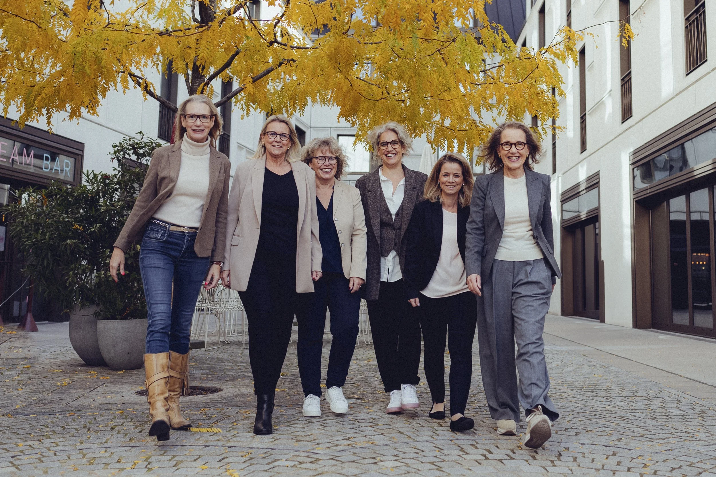 Sechs Frauen in Business-Kleidung gehen auf einer gepflasterten Straße mit Herbstbäumen und modernen Gebäuden.