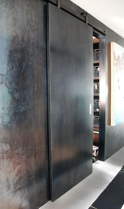 Une porte coulissante noire partiellement ouverte révèle une pièce de rangement avec des étagères en treillis métallique et divers objets à l’intérieur.