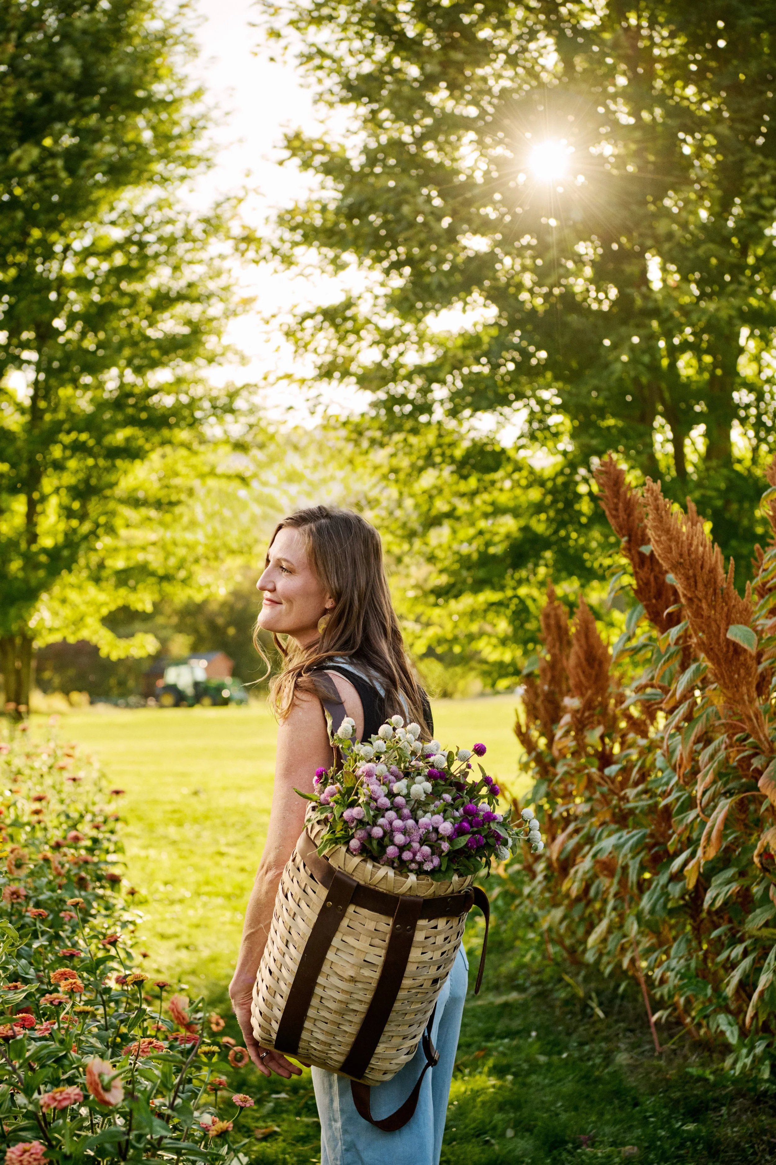 A woman with a woven basket filled with flowers standing outdoors in a lush, green park during sunlight.