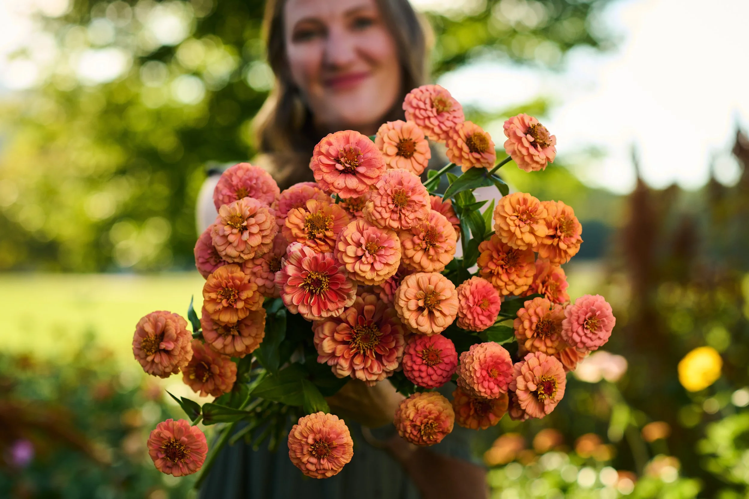 A woman smiling and holding a large bouquet of pink and orange flowers outdoors with greenery in the background.