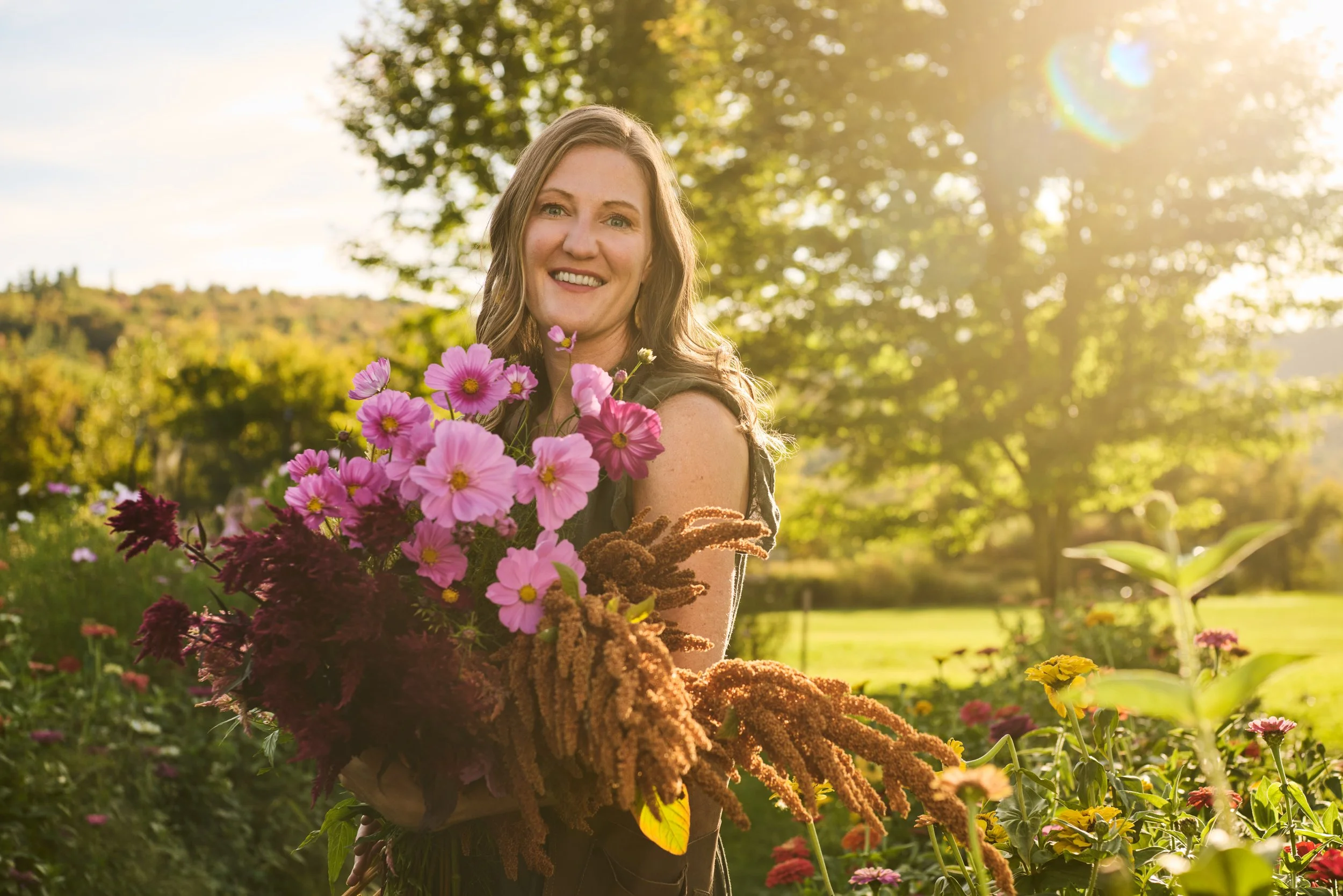 A woman holding a large bouquet of pink and purple flowers in a garden during golden hour sunlight.