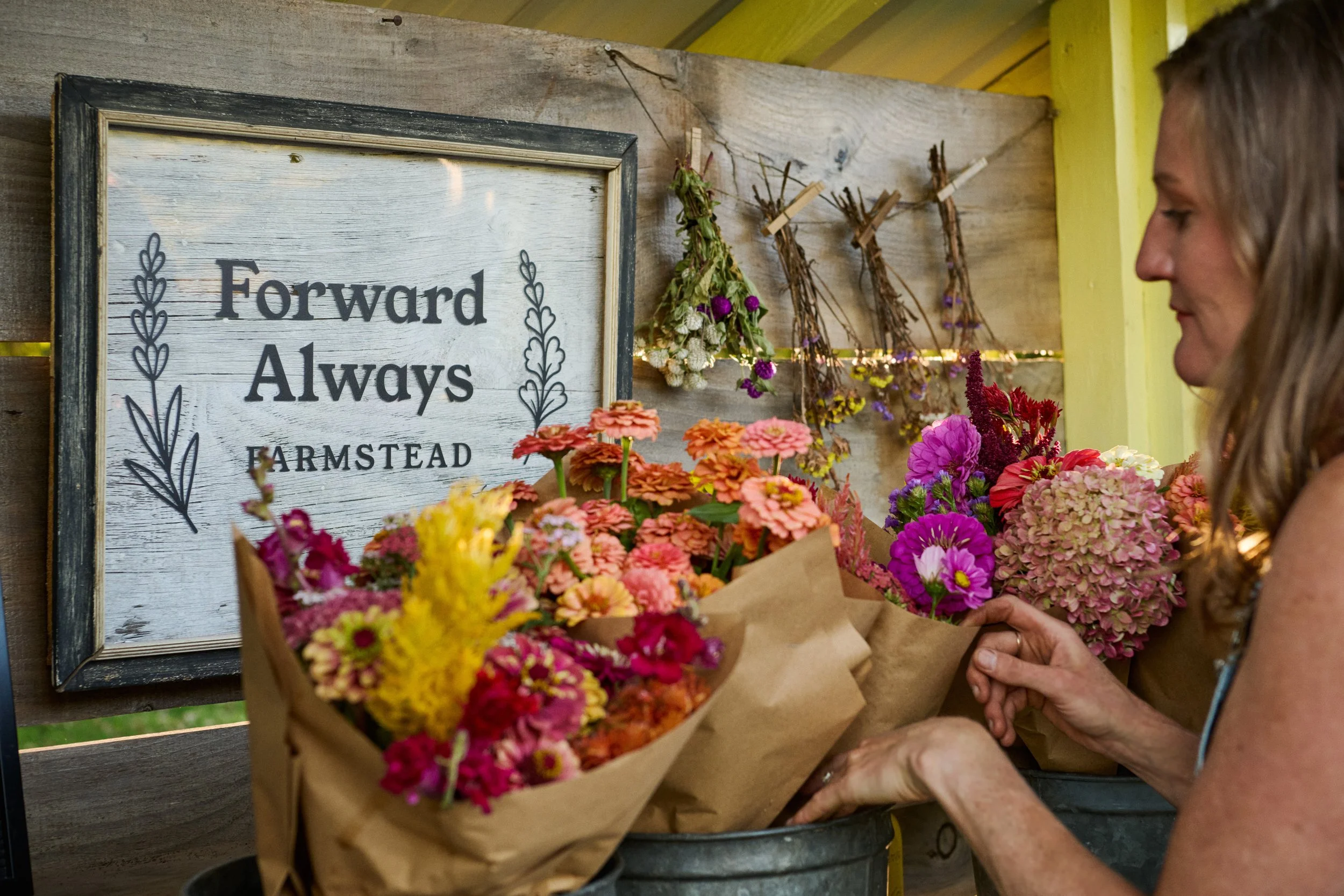 Woman holding a bouquet of pink, purple, and yellow flowers in front of a framed sign that reads 'Forward Always Farmstead,' with dried flower arrangements hanging on a wooden wall.