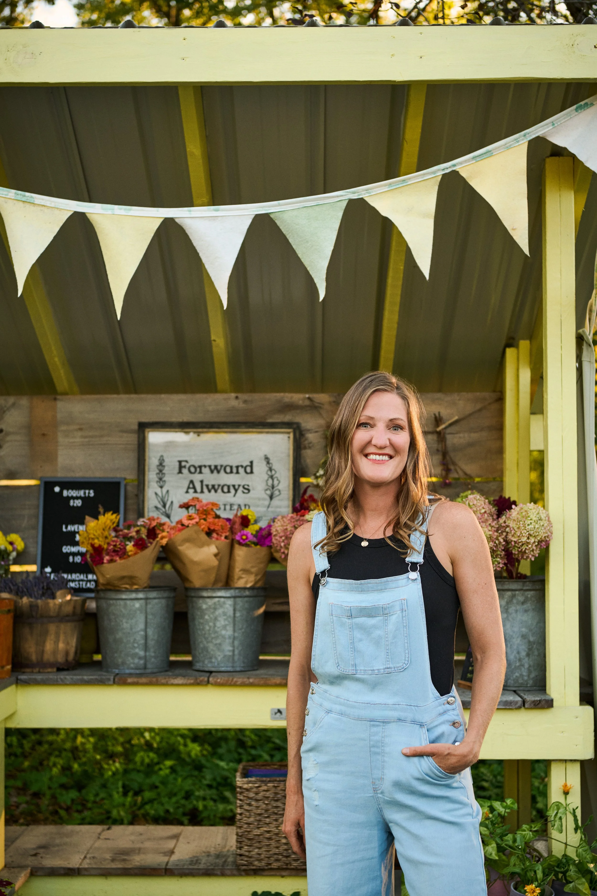 A woman with shoulder-length wavy hair smiling at a flower stand decorated with a banner of light-colored triangular flags.