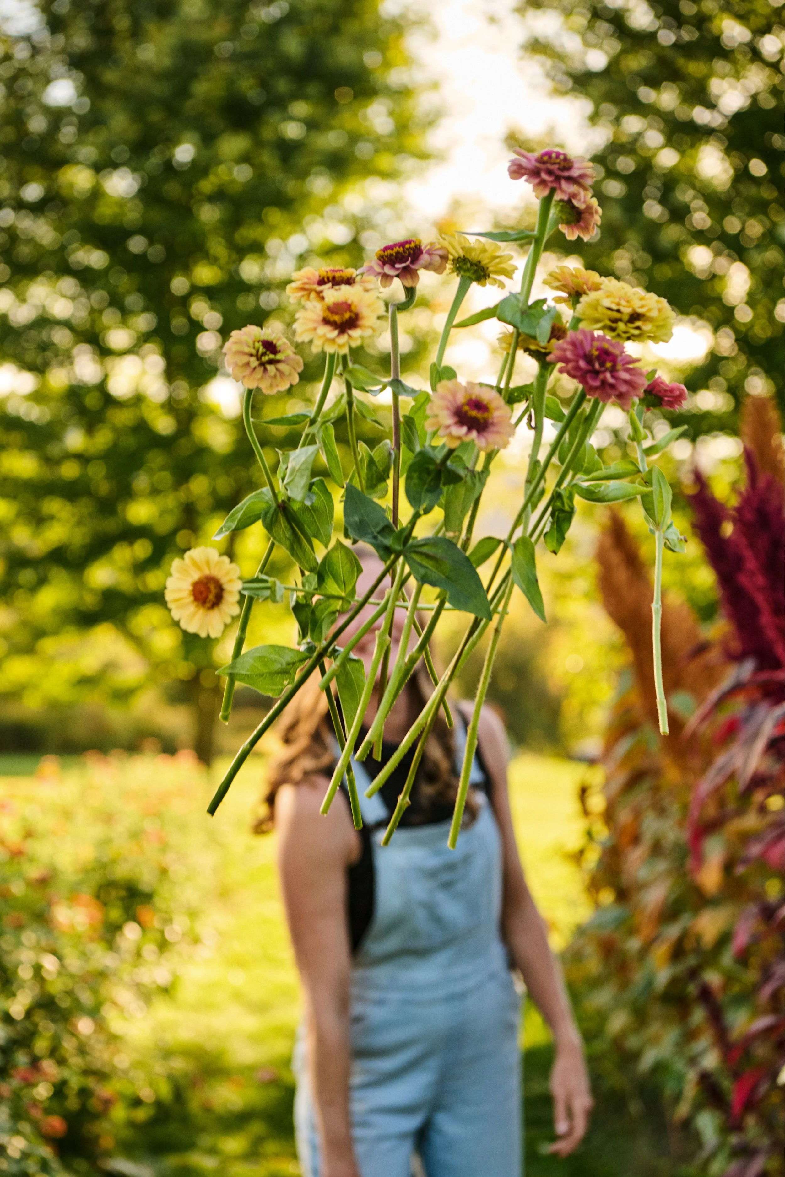 Close-up of a bouquet of colorful flowers in the foreground, with a woman in a light-colored jumpsuit and backpack standing and walking in a garden or park in the background, with lush green trees and bushes, during golden hour sunlight.