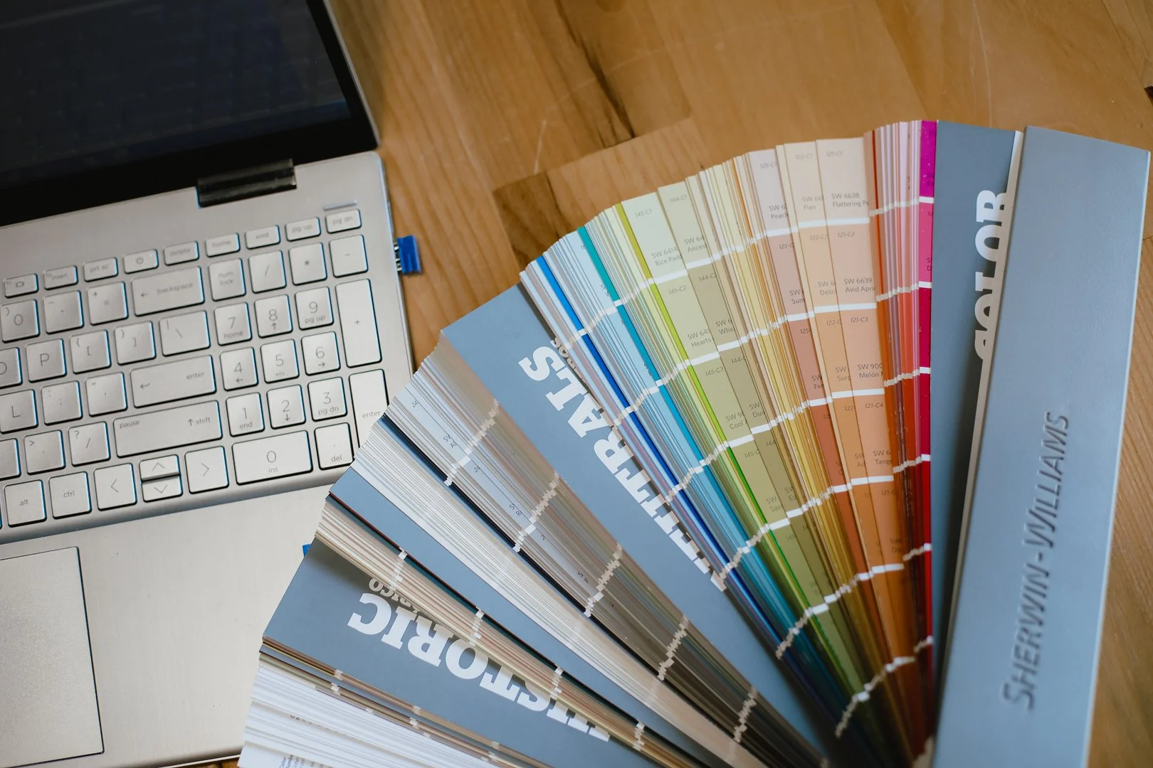 Color sample book fanned out on a wooden table next to a silver laptop keyboard.