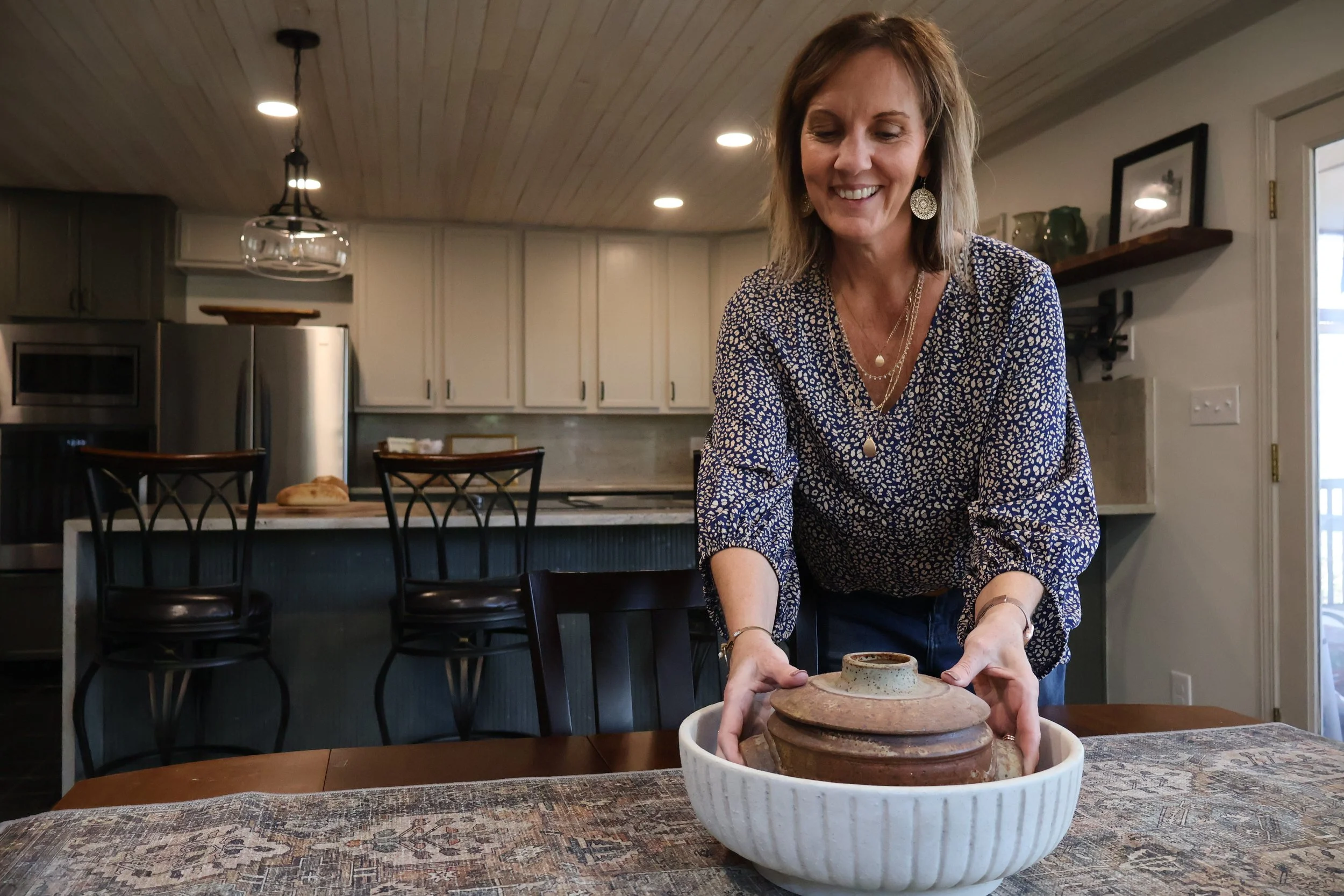 Woman placing a ceramic pot into a white bowl on a dining table in a kitchen.