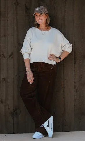 Woman standing in front of wooden wall, wearing a white sweater, dark pants, white sneakers, and a patterned hat.