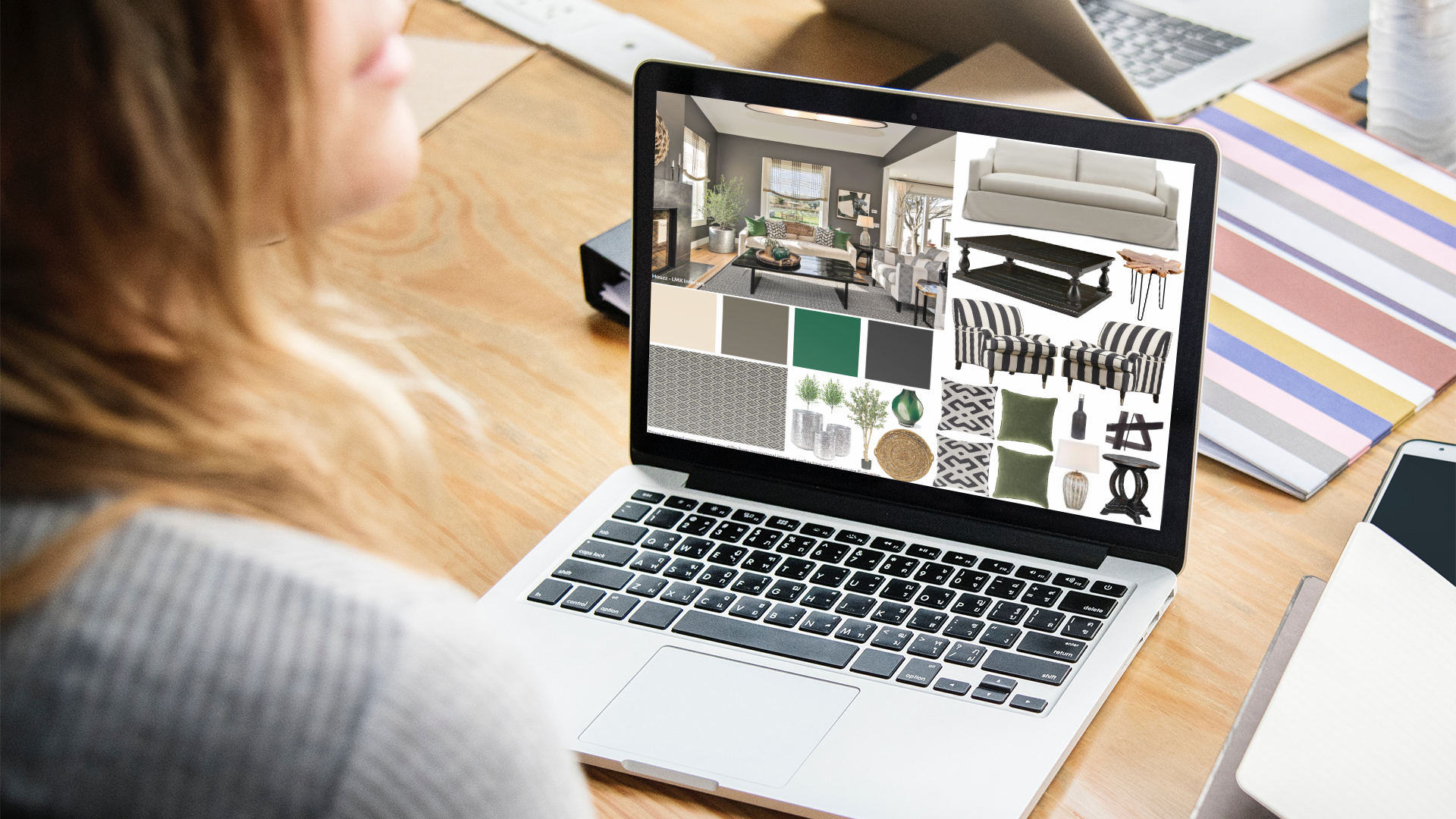 A person working on a laptop with interior design images and fabric swatches on the screen. The workspace includes a wooden table, other laptops, notebooks, and files.
