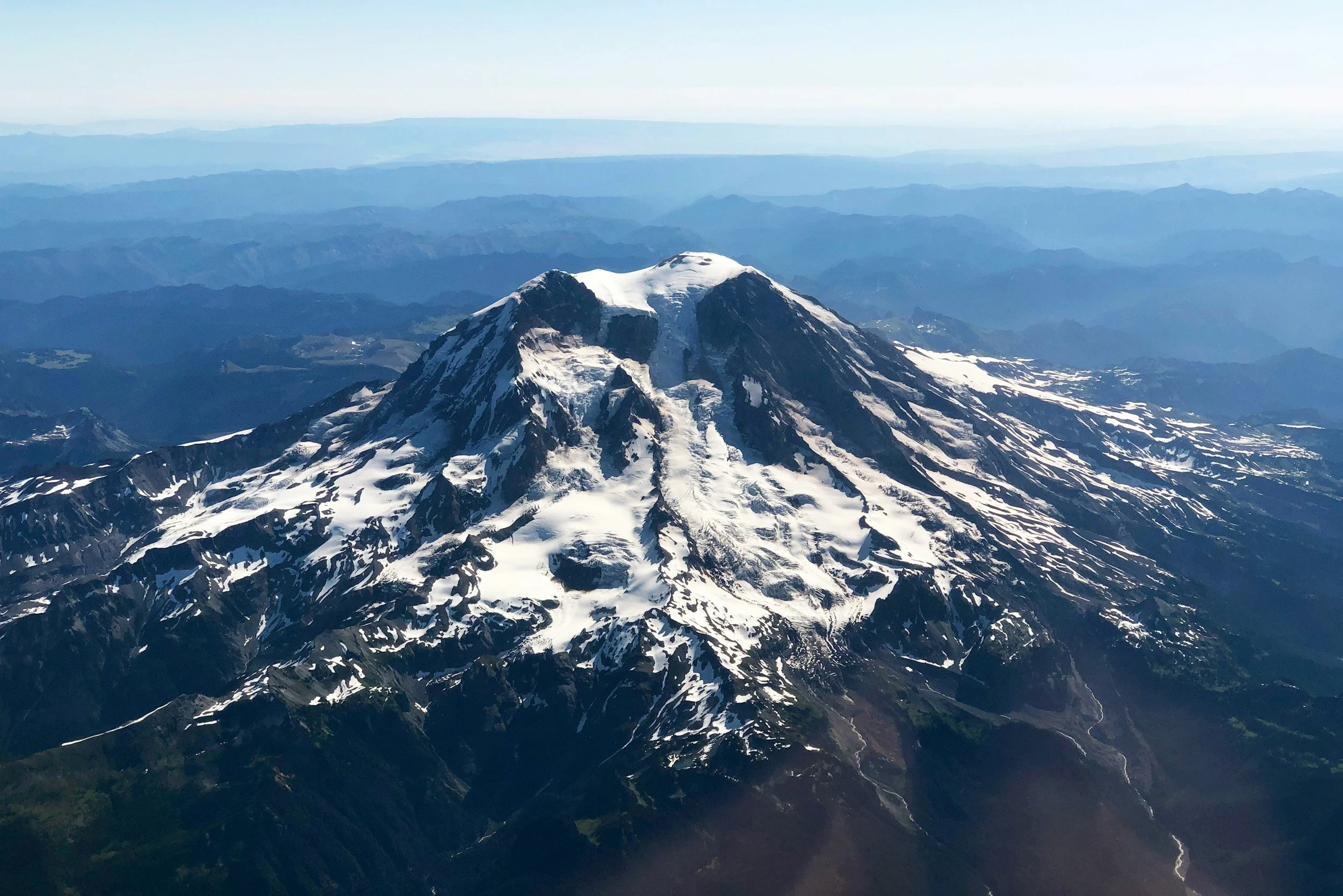 A snow-capped mountain with rugged peaks and slopes, surrounded by a landscape of rolling hills and distant ranges under a clear sky.