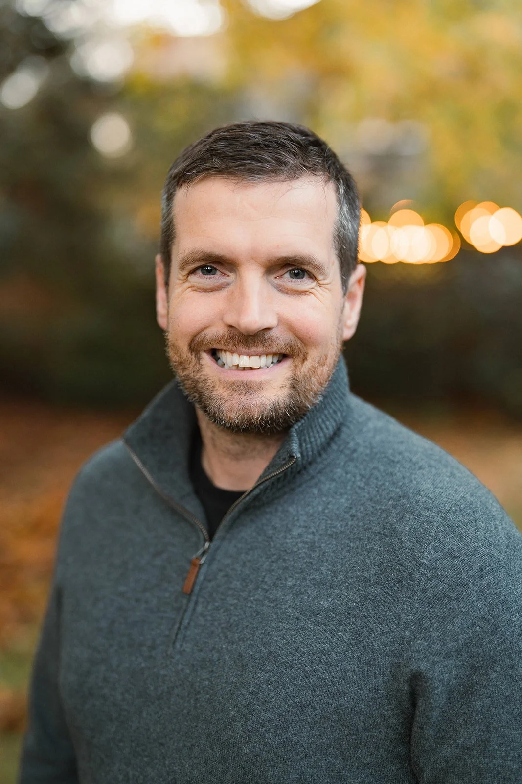 Headshot of a smiling man with short dark hair, beard, wearing a gray sweater, outdoors with autumn trees in the background.