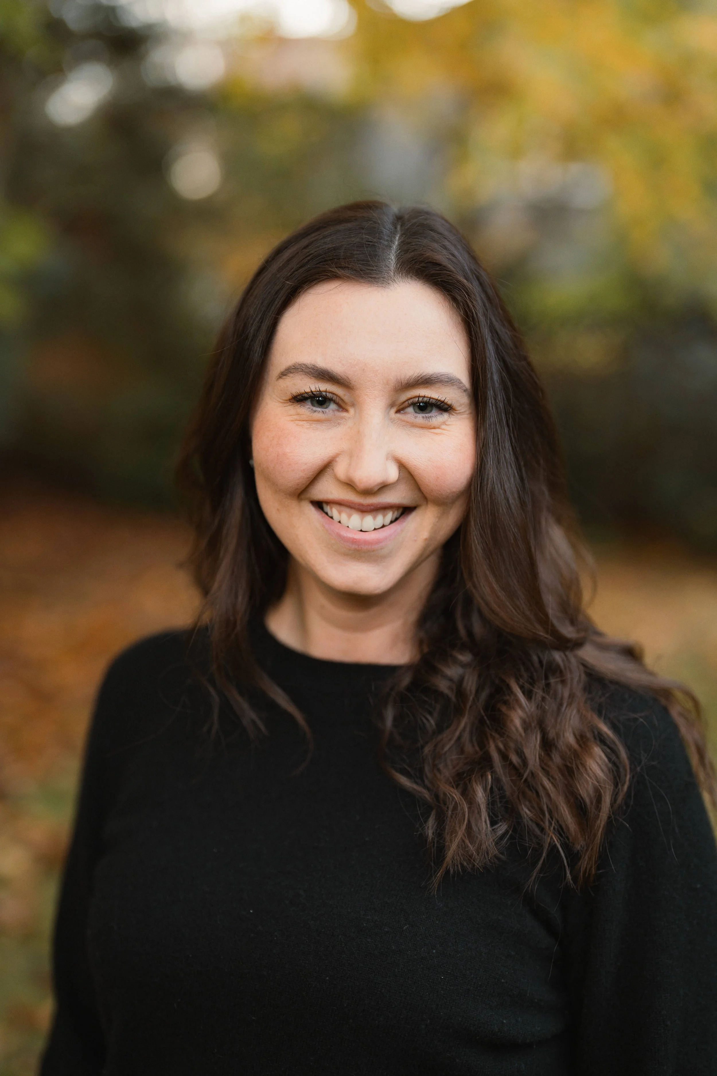 A woman with long wavy brown hair and blue eyes smiling outdoors with a blurred background of autumn trees.