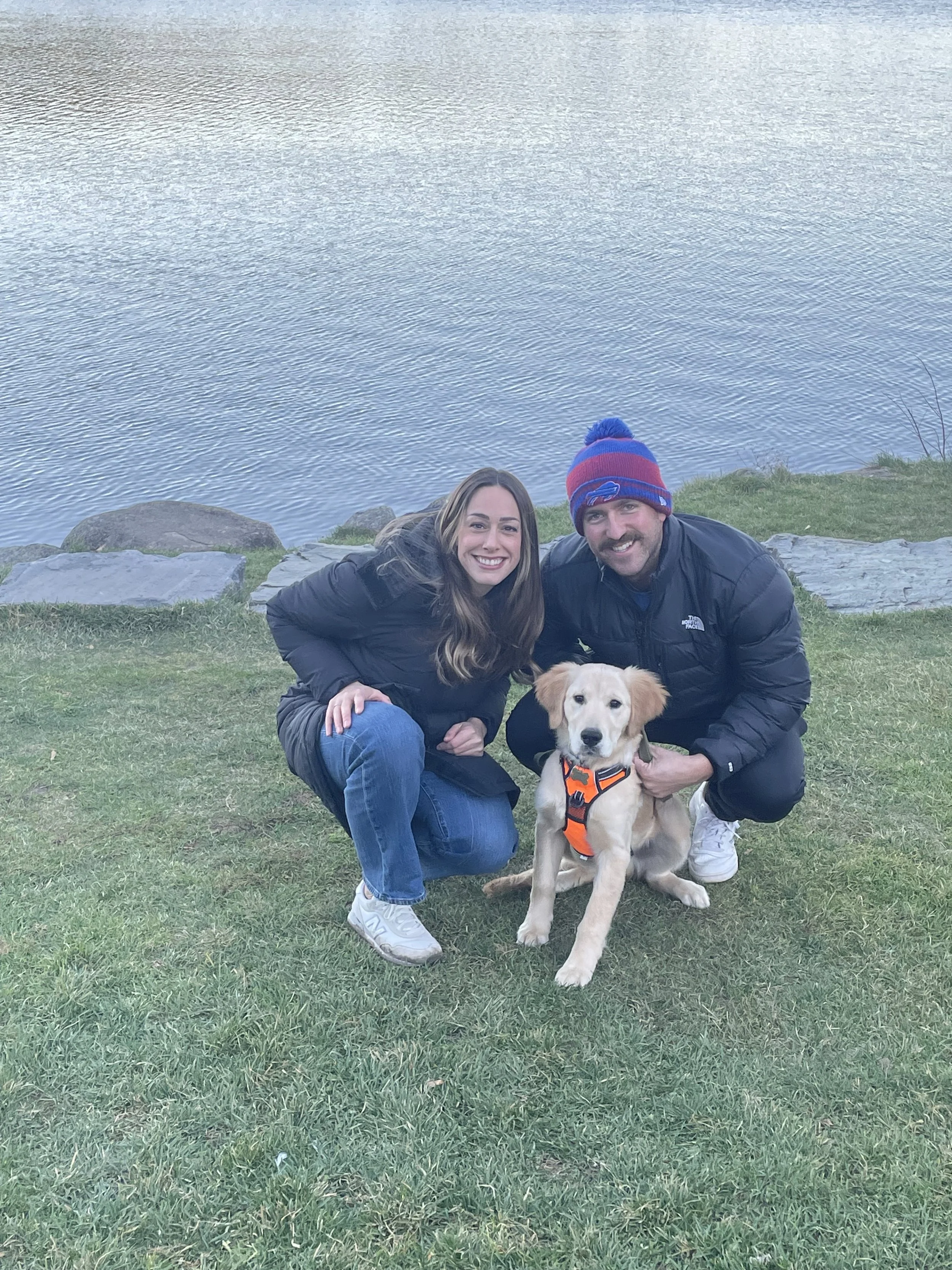 A woman, a man wearing a red and blue beanie, and a golden retriever puppy in an orange harness are outdoors near a body of water with rocks and grass.