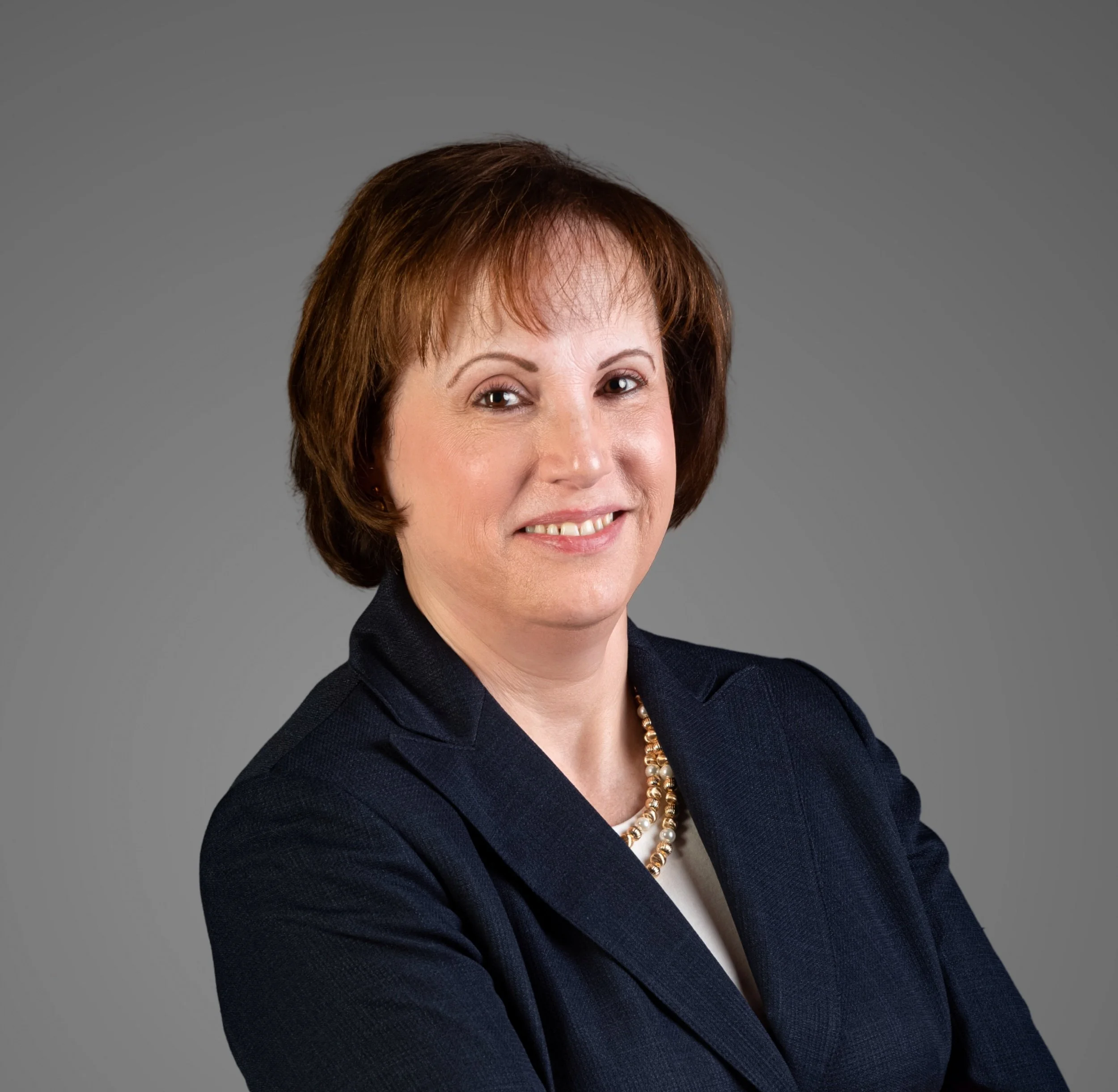 A professional woman with short brown hair, wearing a dark blazer and a pearl necklace, smiling with arms crossed against a gray background.