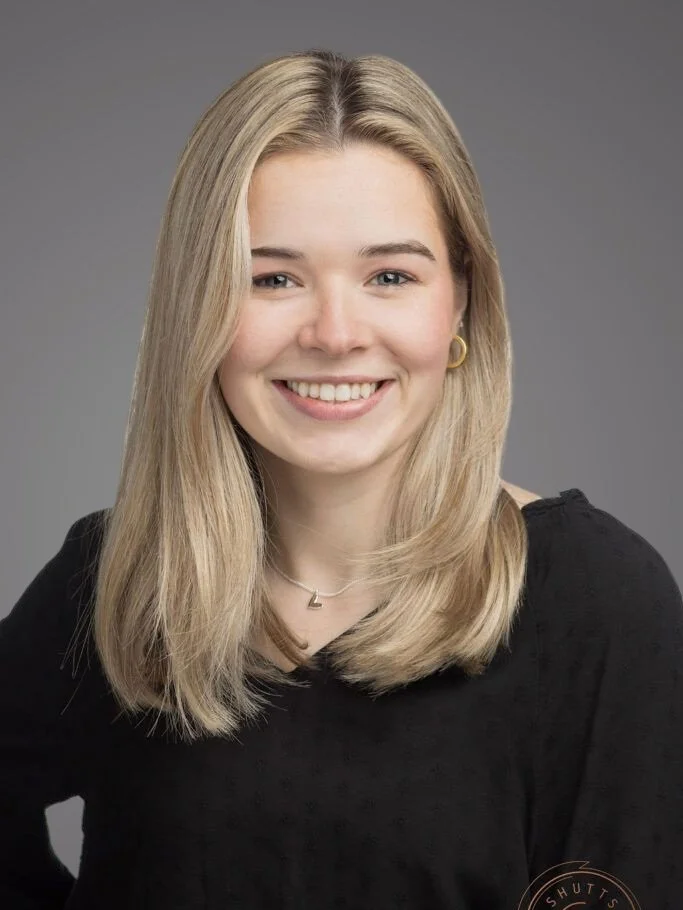 A young woman with blonde hair, smiling, wearing a black top, gold hoop earrings, and a delicate necklace with a small pendant, against a plain grey background.