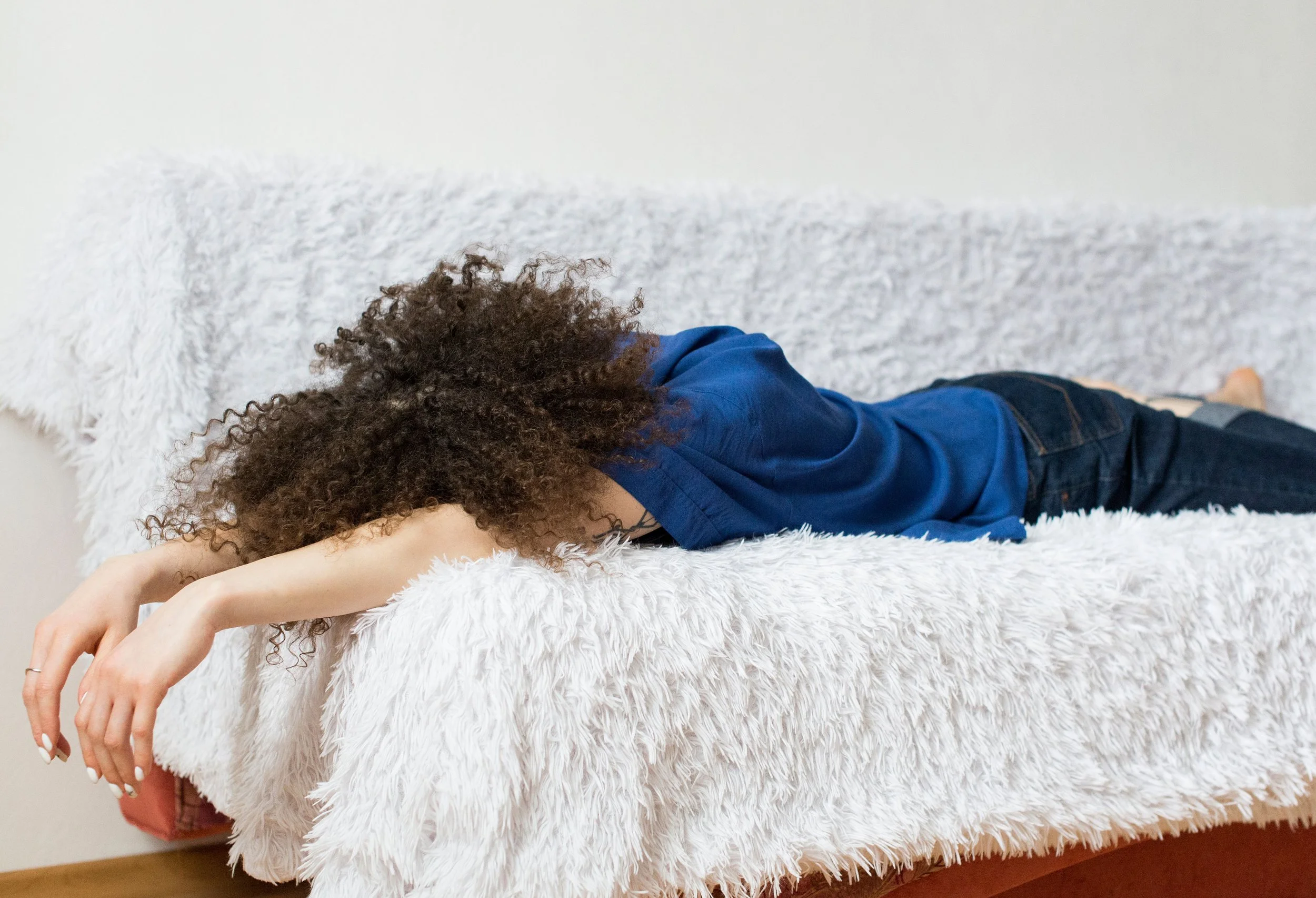Person with curly hair and blue t shirt and jeans lying facedown on white fluffy couch with arms hanging off the side of the couch