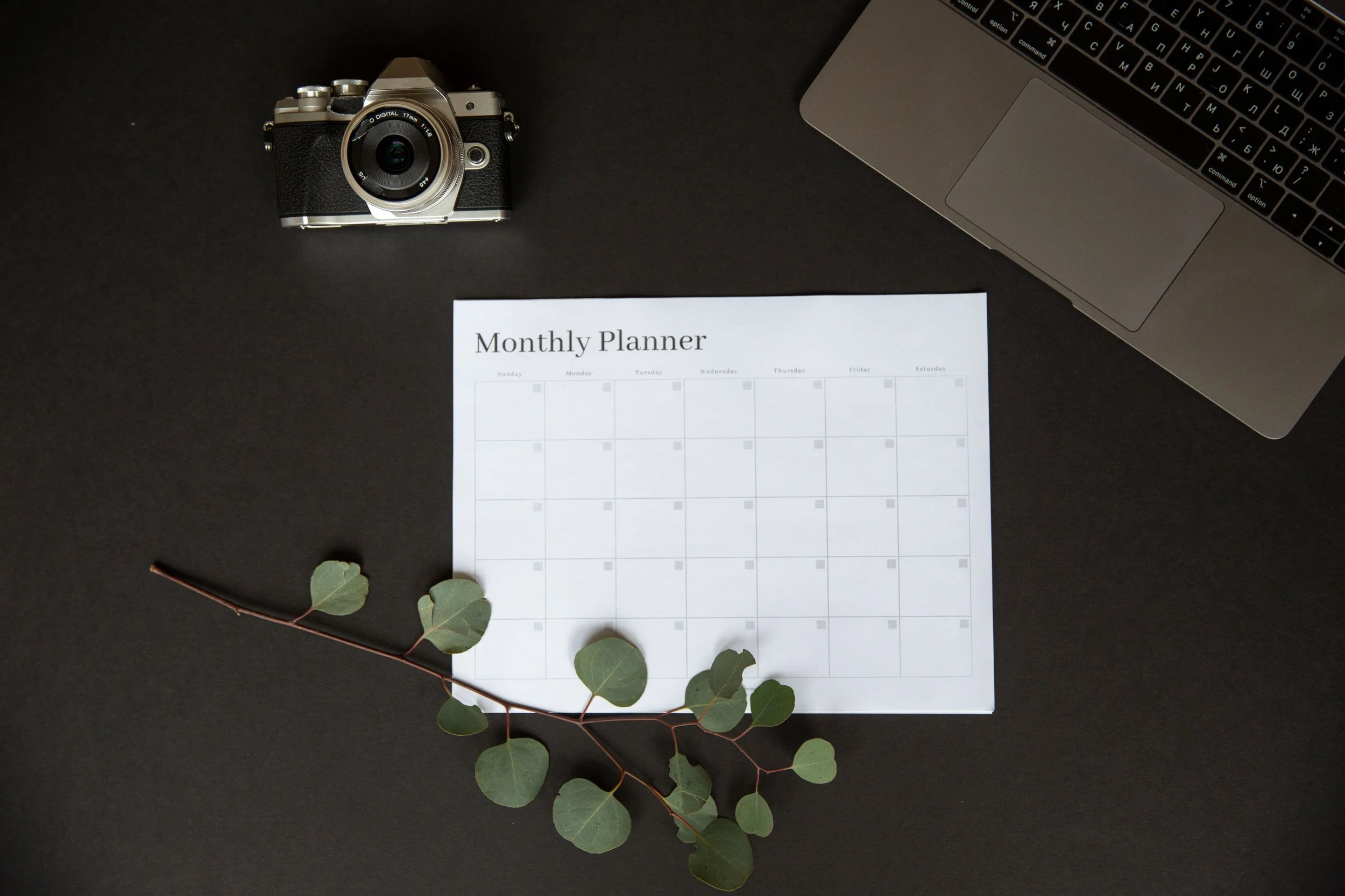 empty monthly planner page on black desk in centre with branch with green leaves layered on top and camera in top left and computer in top right of photo