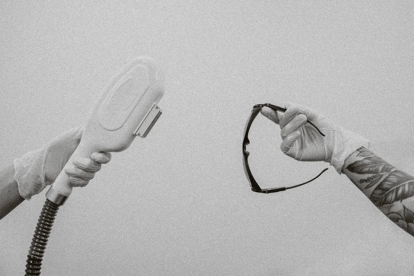 Two hands, one holding a laser and the other holding a pair of safety glasses with a tattooed arm, both wearing gloves, against a plain background.