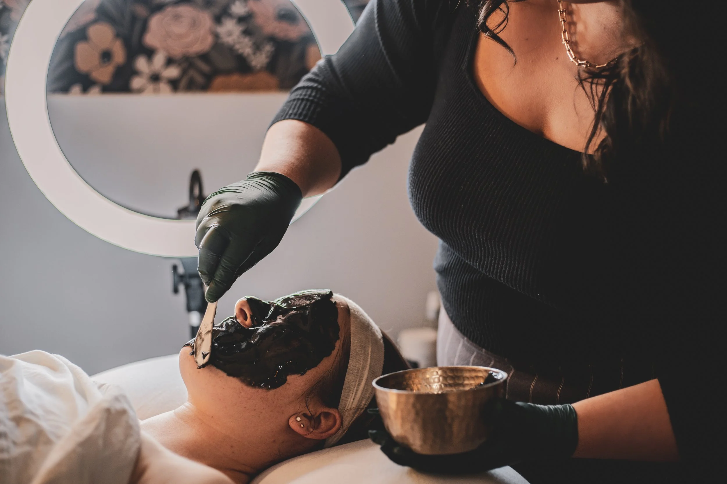 A woman lying face up receiving a facial mask at a spa or skincare clinic, while a skincare professional applies a black facial mask with a spoon.