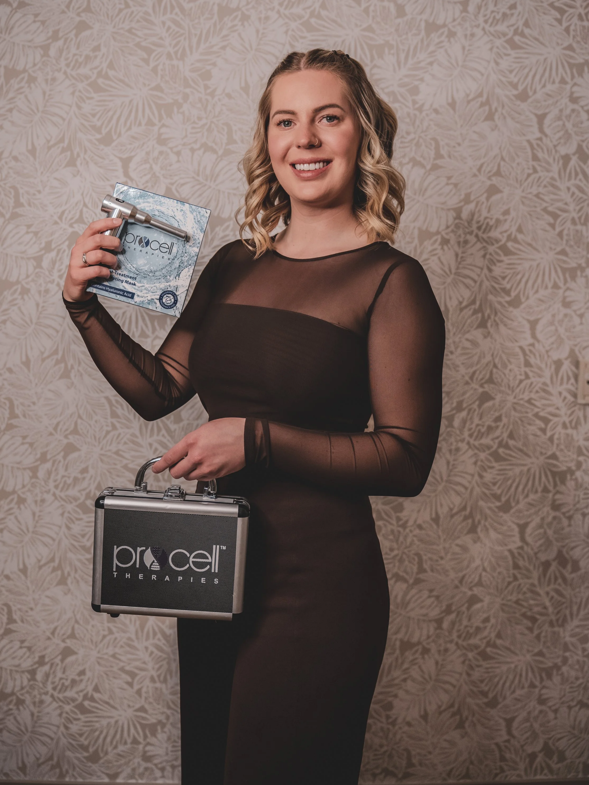 Cheyenne with blonde wavy hair and a nose ring holding a box of ProCell therapies skincare product in one hand and a black briefcase with ProCell Therapies logo in the other, standing against a patterned beige wall.
