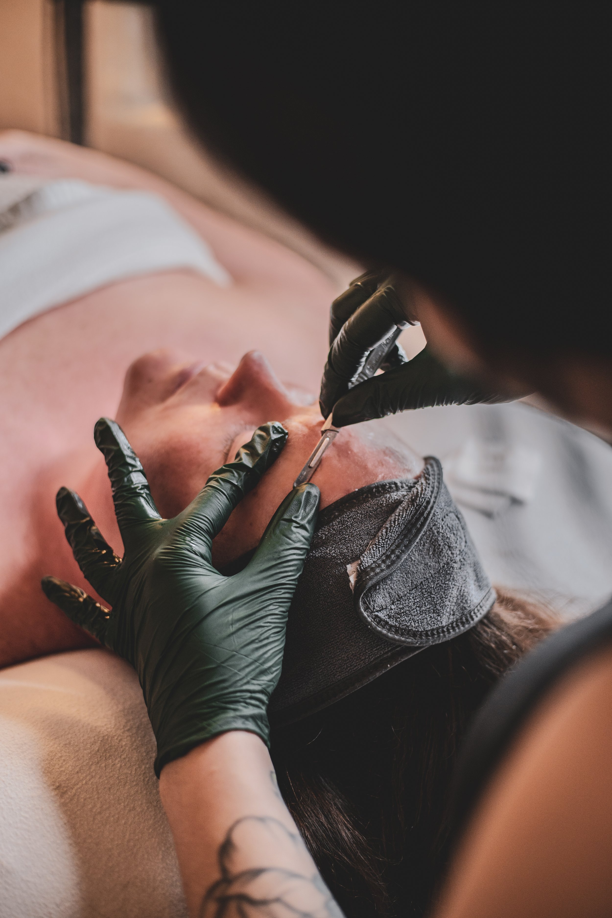 A person receiving a custom facial, laying down on a salon bed. The technician wears black gloves and dermaplanes the client's skin.