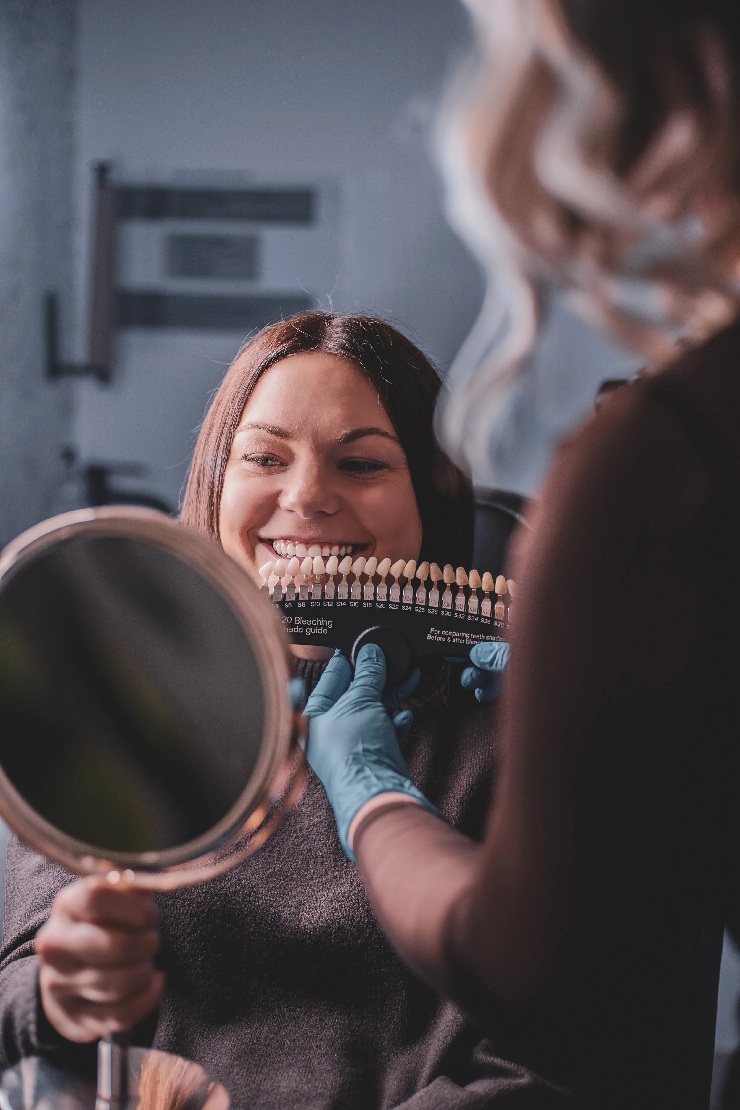 A person holds examples of shades for teeth whitening next to a smiling client looking into a mirror in a salon
