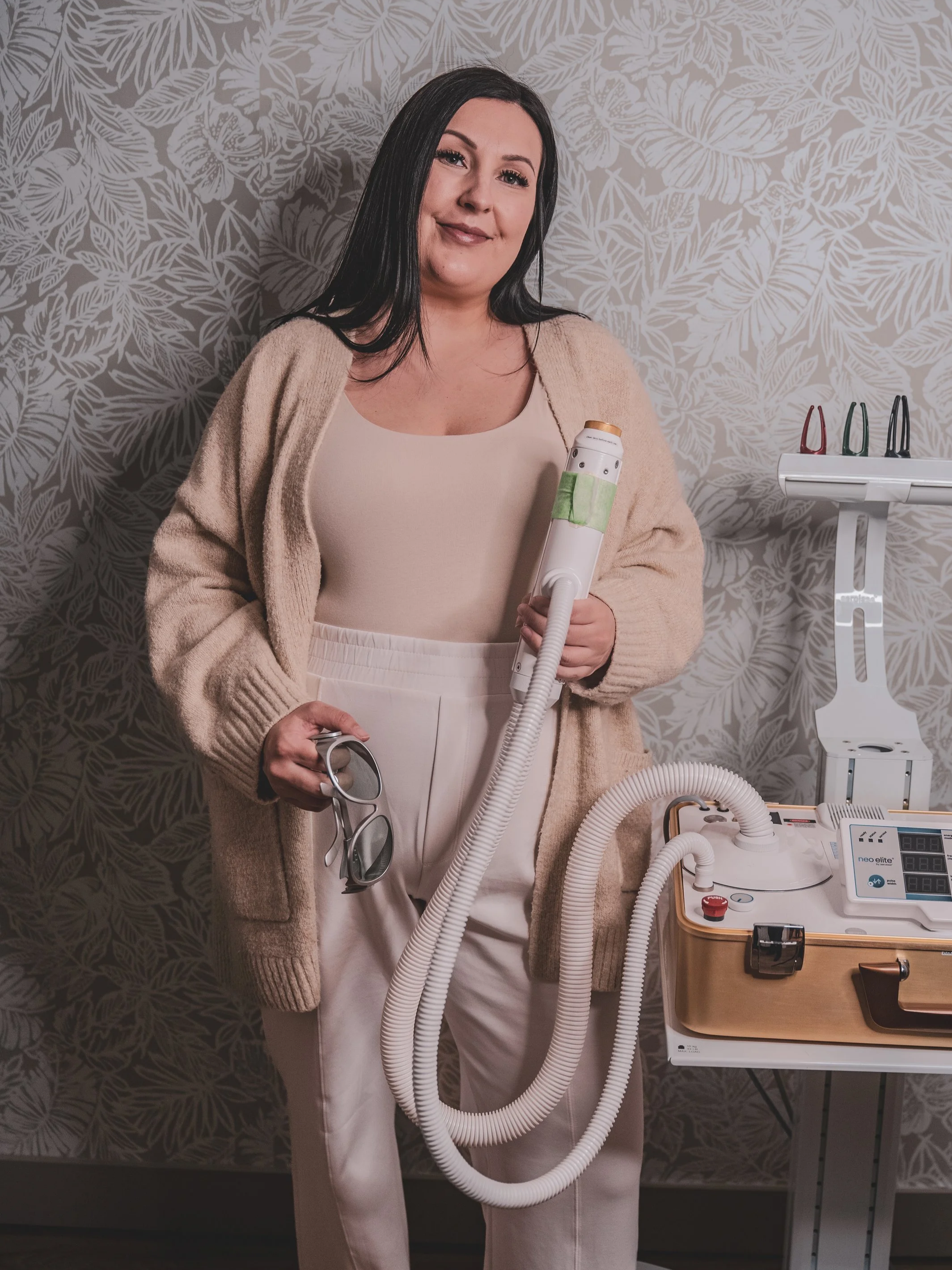 Nicole holding a laser device, standing in a room with a patterned wallpaper background.