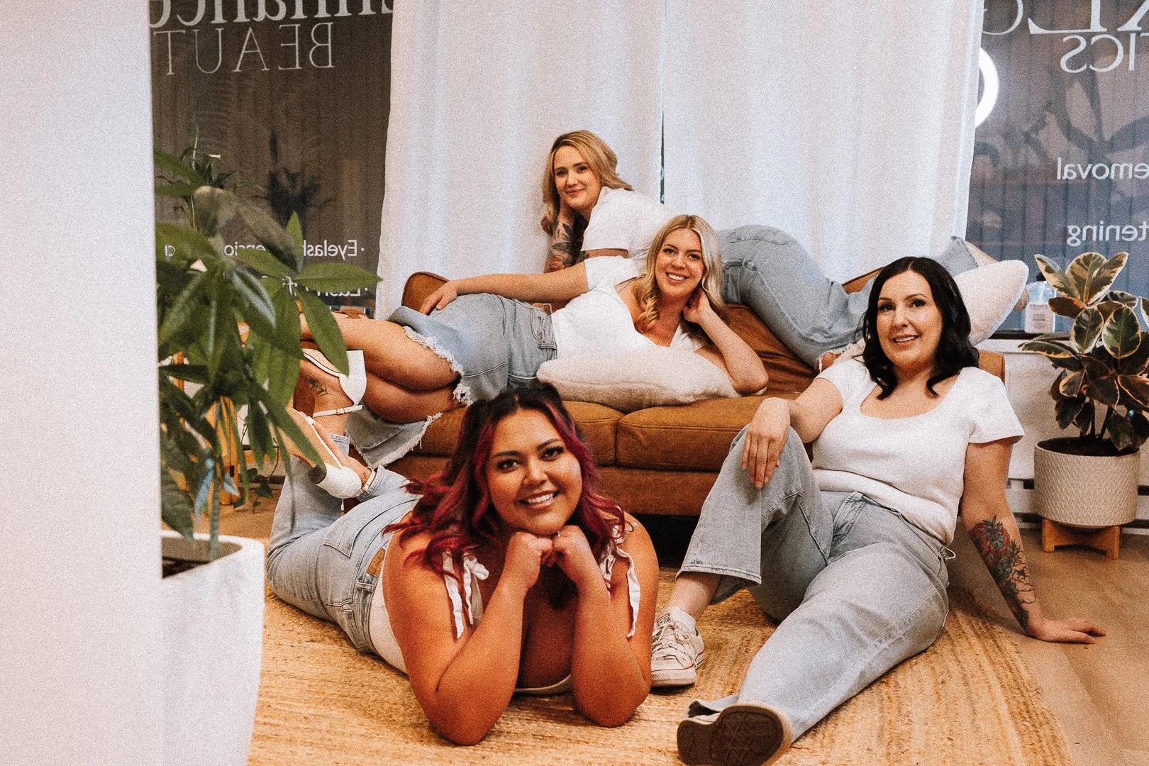 Five women with diverse hair colors and styles relaxing together indoors on a brown couch and a rug, smiling and looking at the camera.