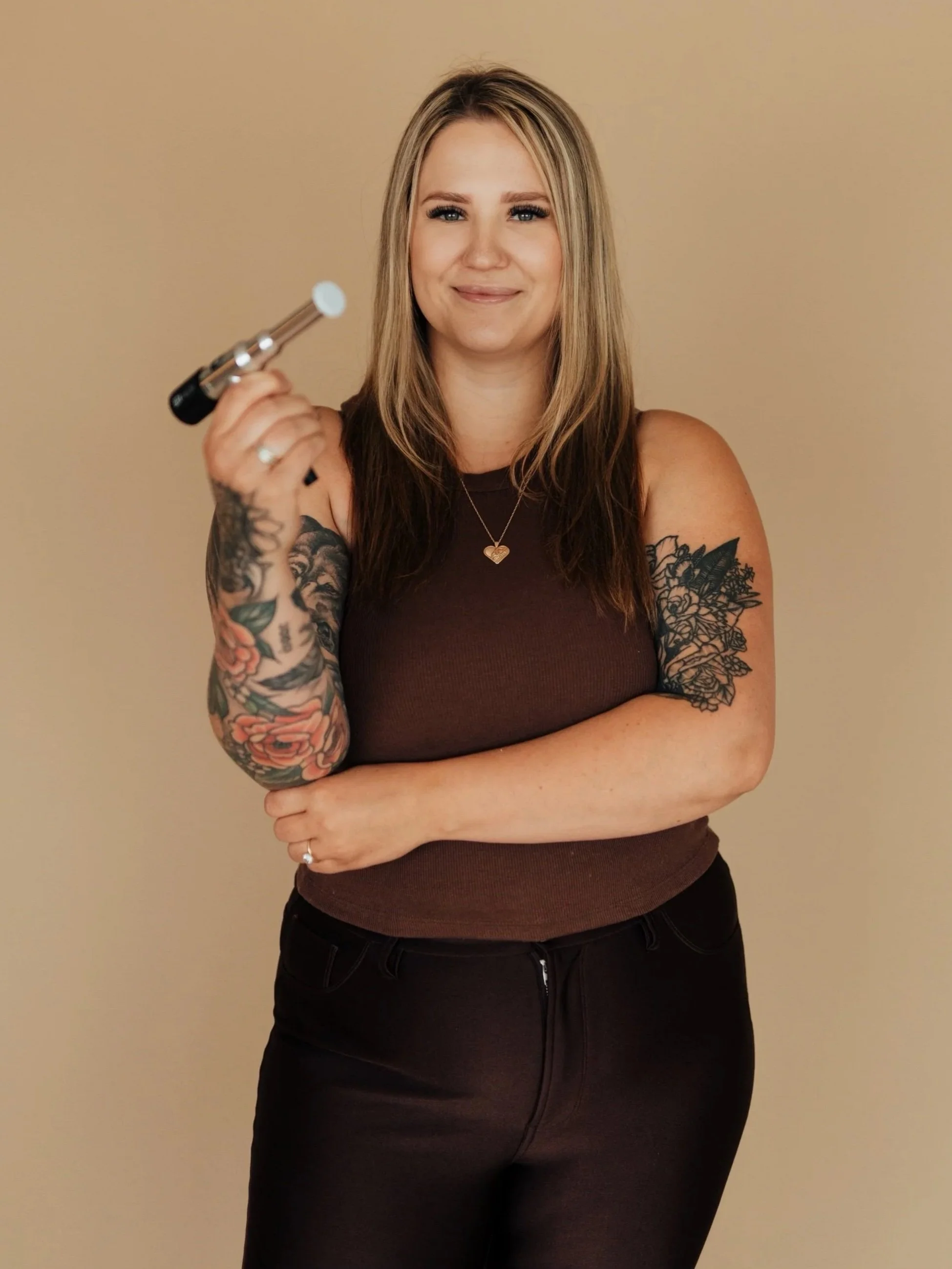 Cleo with blonde hair, tattoos on both arms, wearing a brown sleeveless top and black pants, holding a ProCell gun, smiling, standing against a beige background.